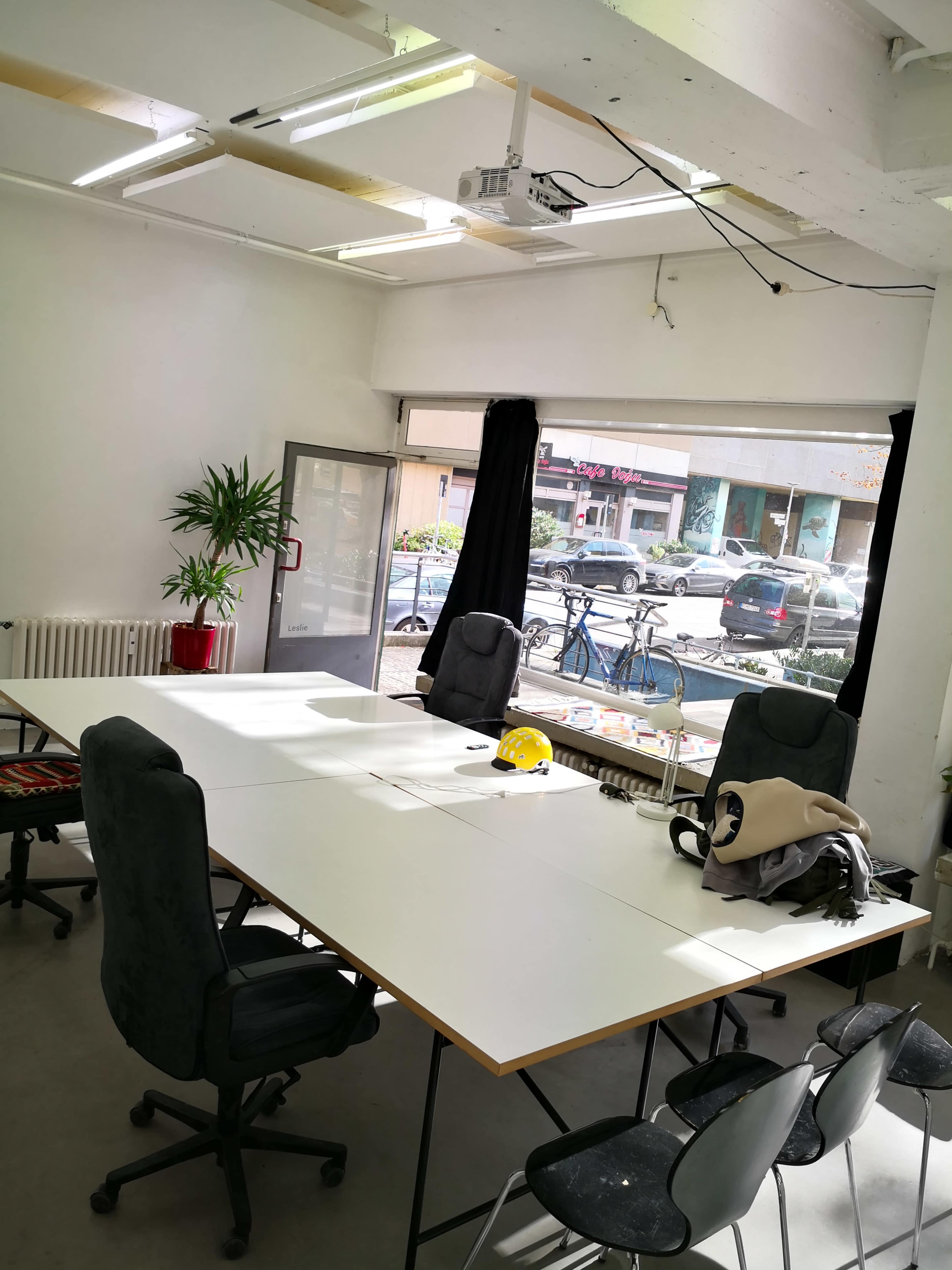 A brightly lit meeting room features a large table surrounded by chairs, with a projector mounted on the ceiling and a window showing street views outside.