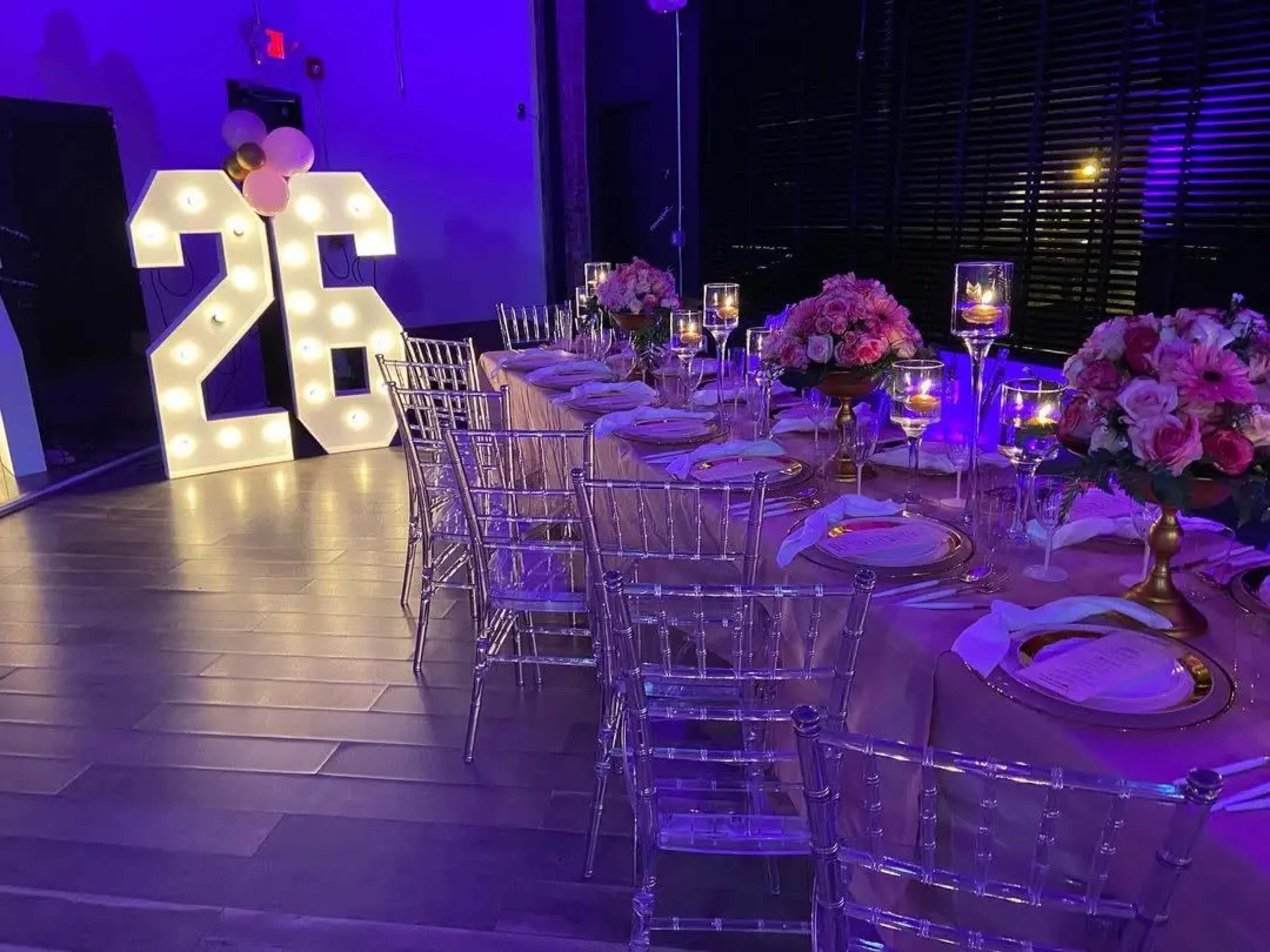 The image shows a decorated dining area featuring a long table with elegant place settings, floral centerpieces, and illuminated numbers "26" in the background.