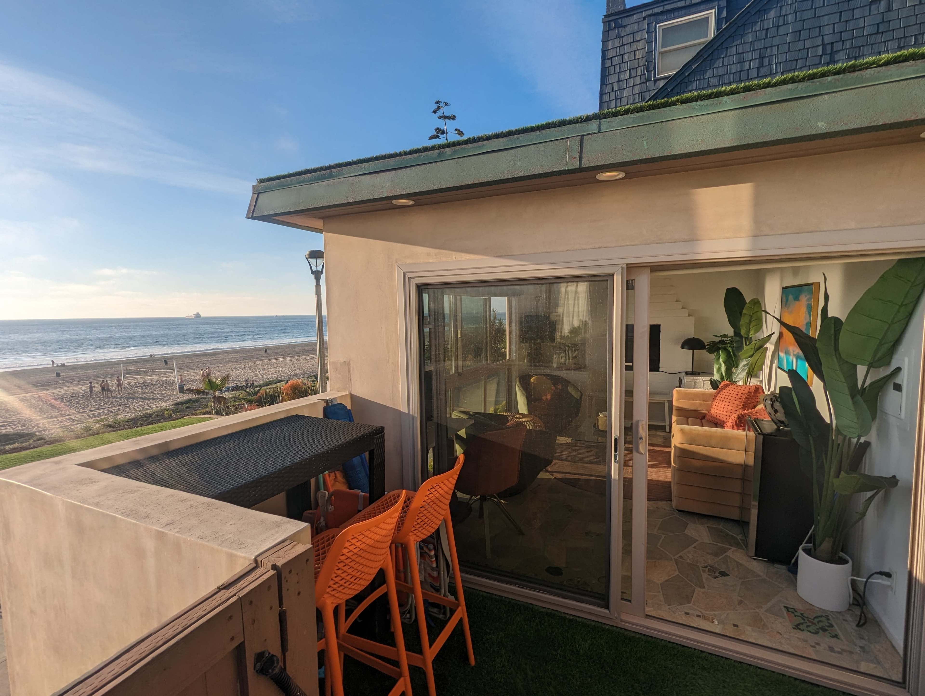 A coastal view from a balcony with orange bar stools overlooking a beach, and a bright living room visible through the sliding glass door.