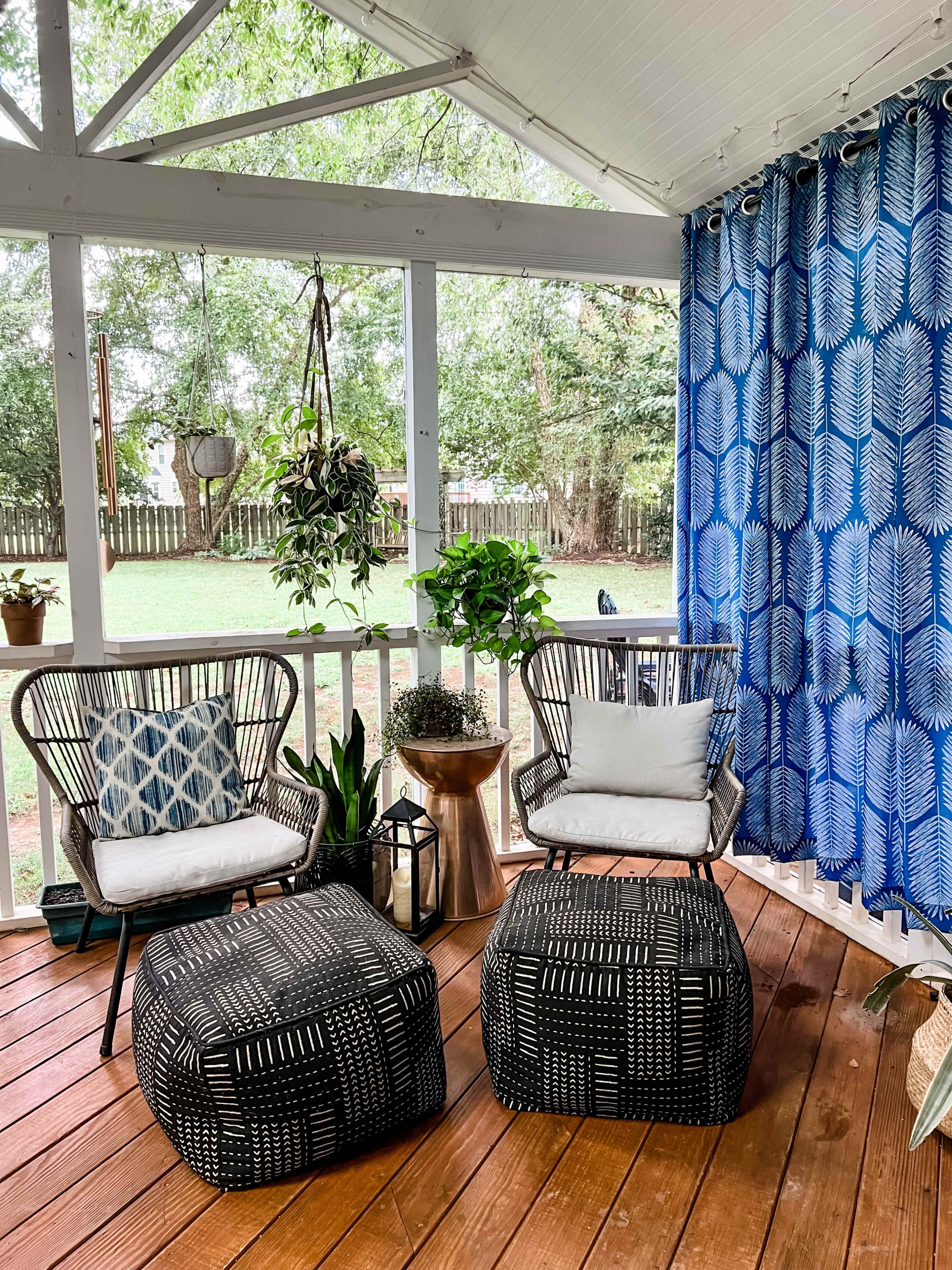 A covered outdoor space with two black patterned ottomans, two chairs with cushions, a small table, and plants arranged on a wooden deck.