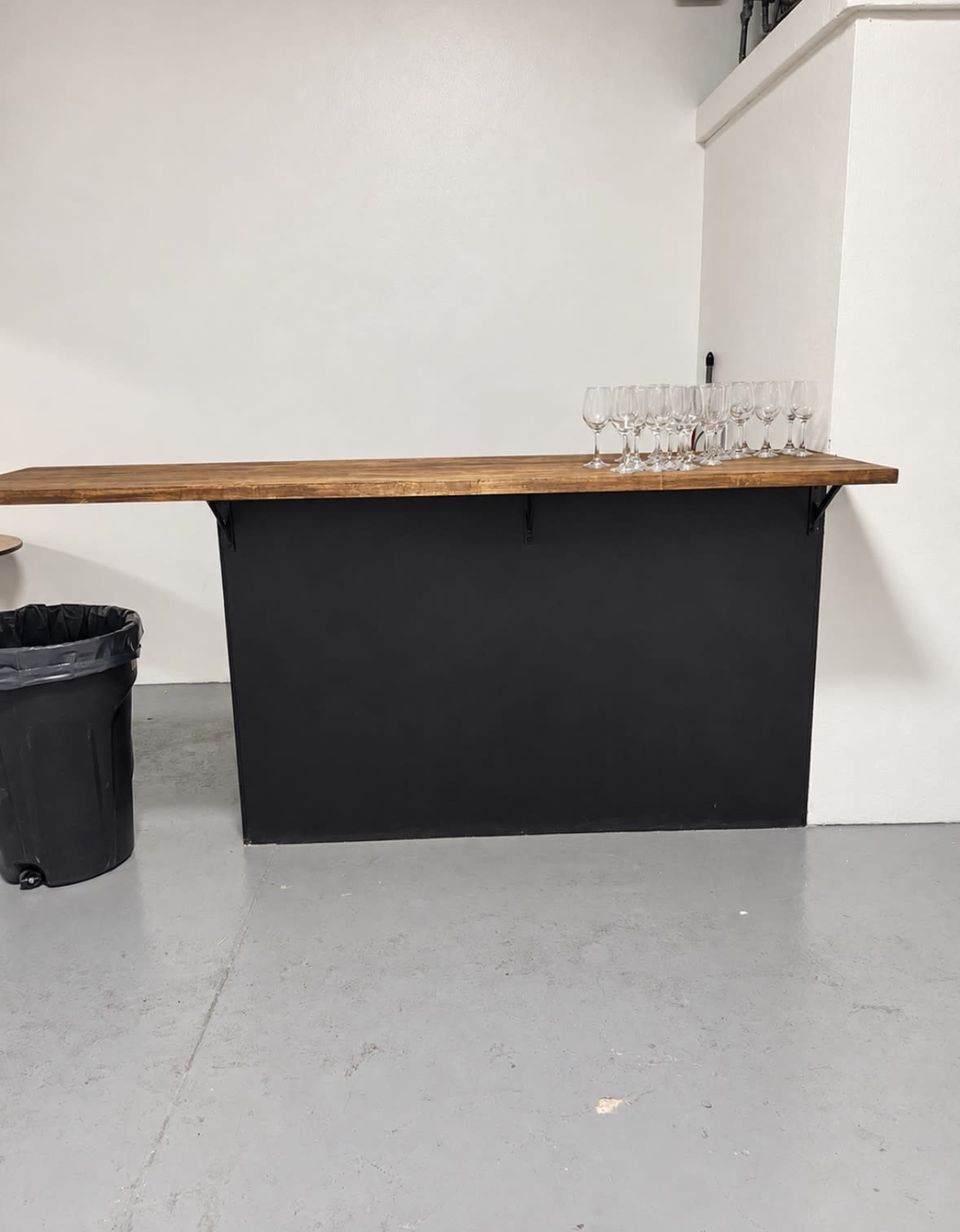 The image shows a black countertop with a wooden ledge and a row of clean glasses on top, next to a trash bin in a plain room.