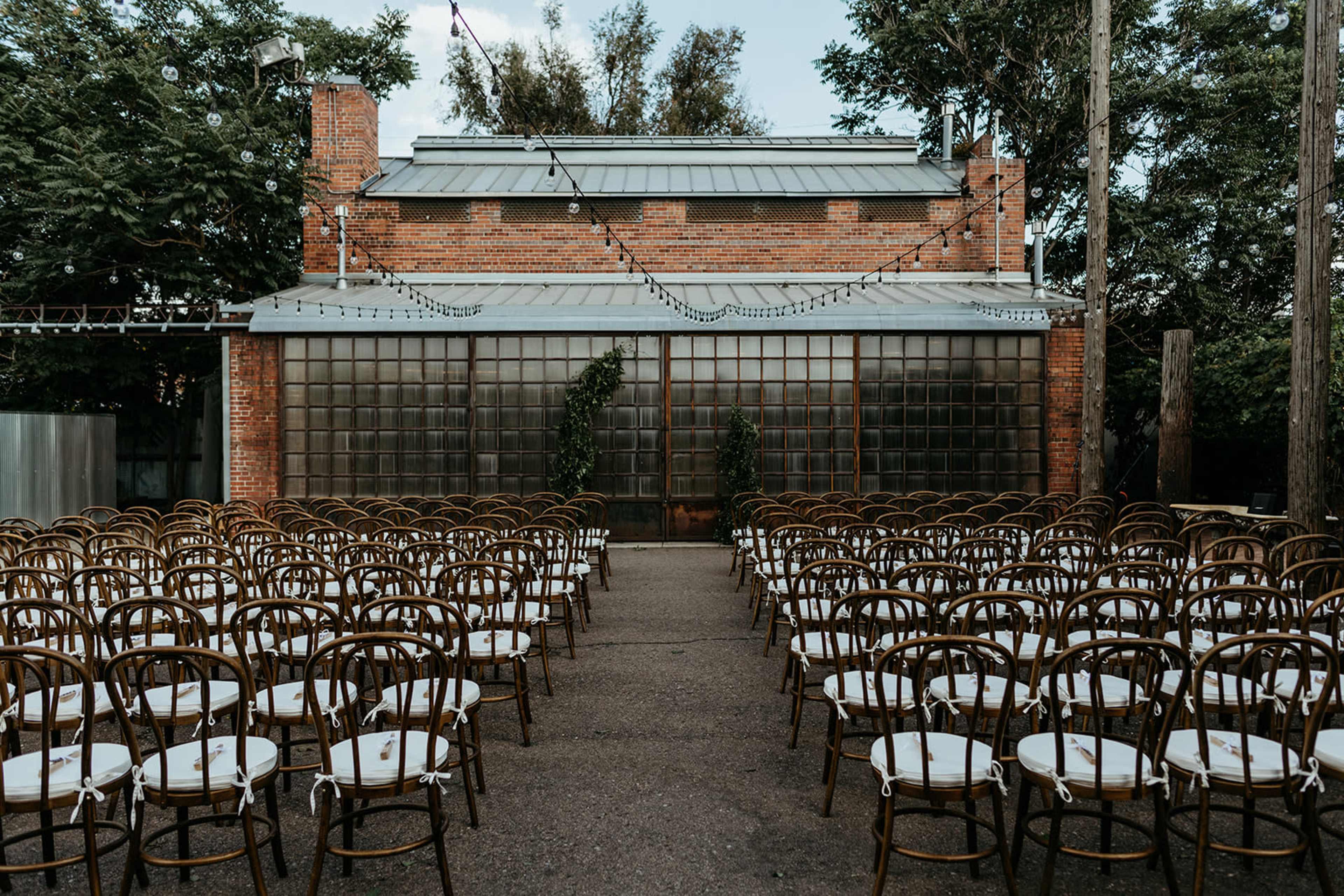 A setup for an outdoor event with rows of wooden chairs arranged in front of a brick building featuring large glass windows.