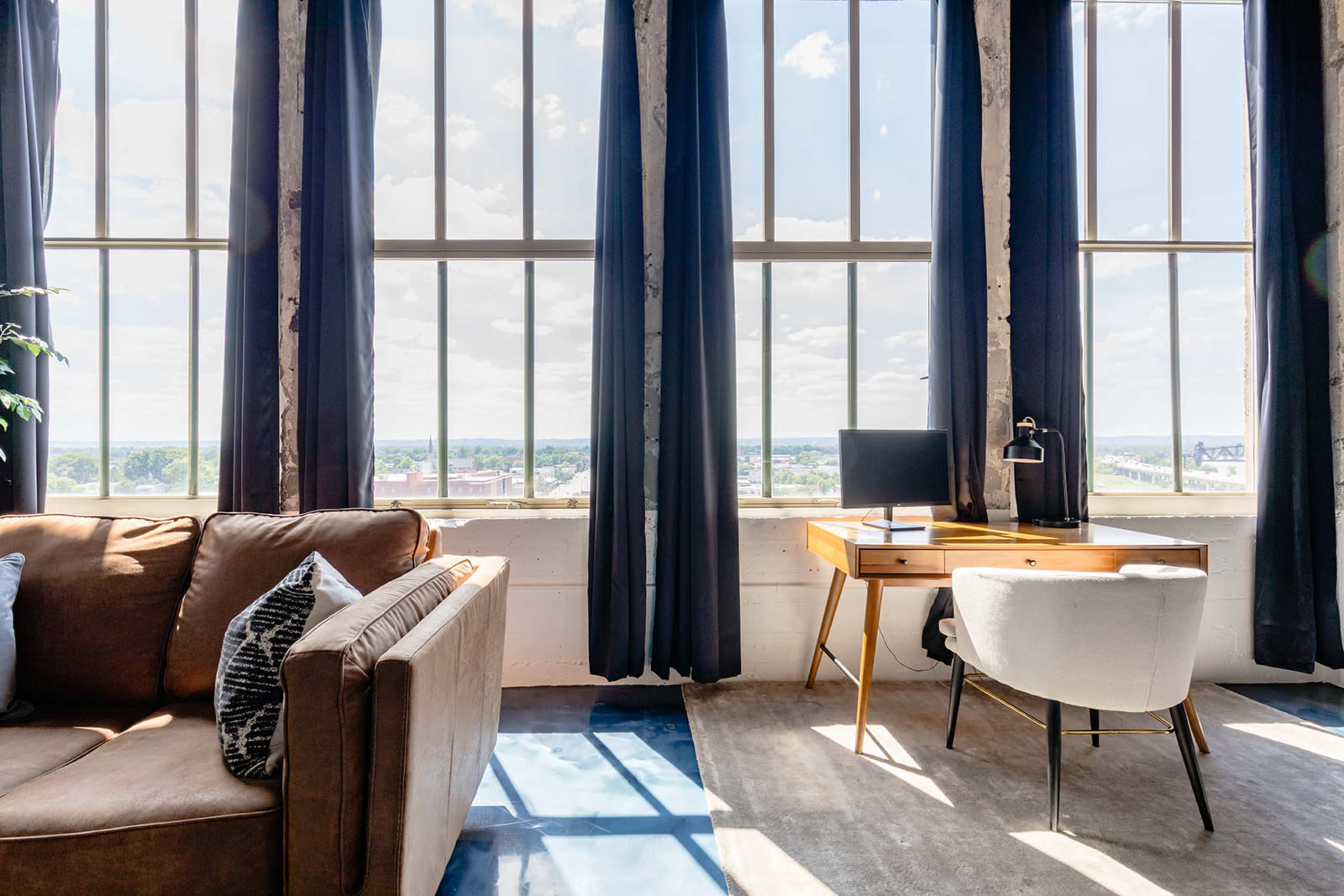 The image shows a spacious room with large windows, a sofa, a desk with a computer, and a scenic view of the outdoors.