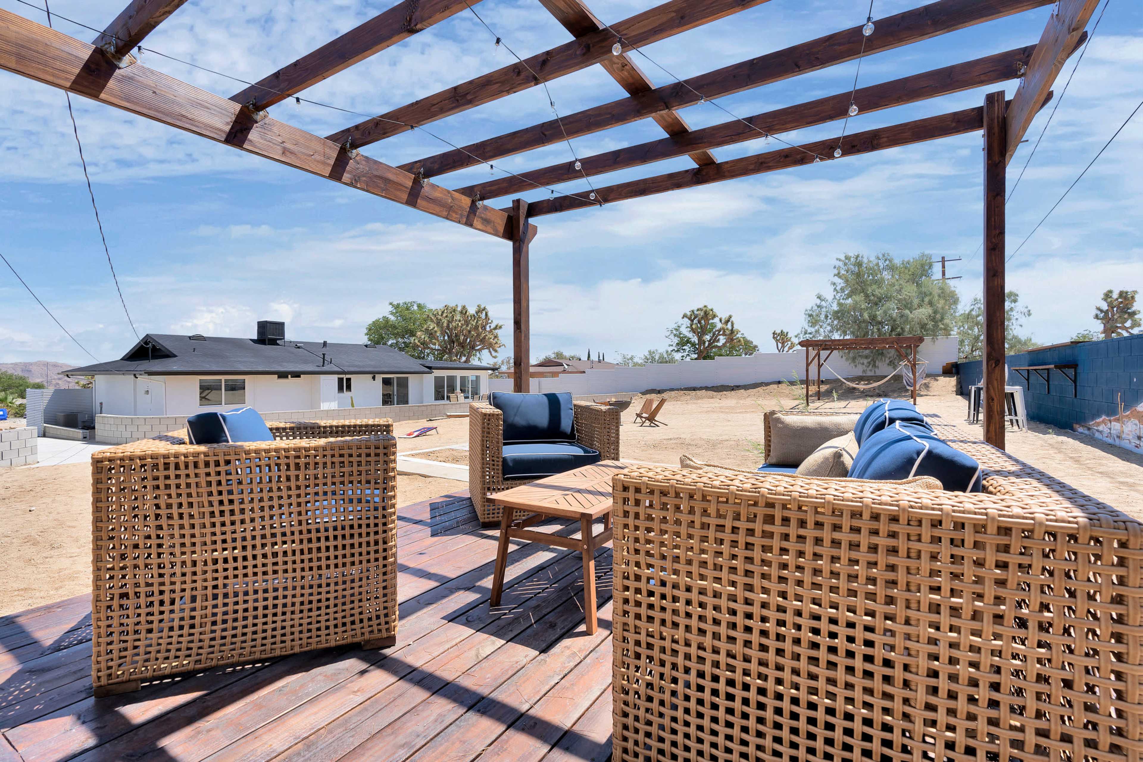 A wooden pergola with string lights frames a seating area featuring wicker chairs on a deck overlooking a barren backyard.