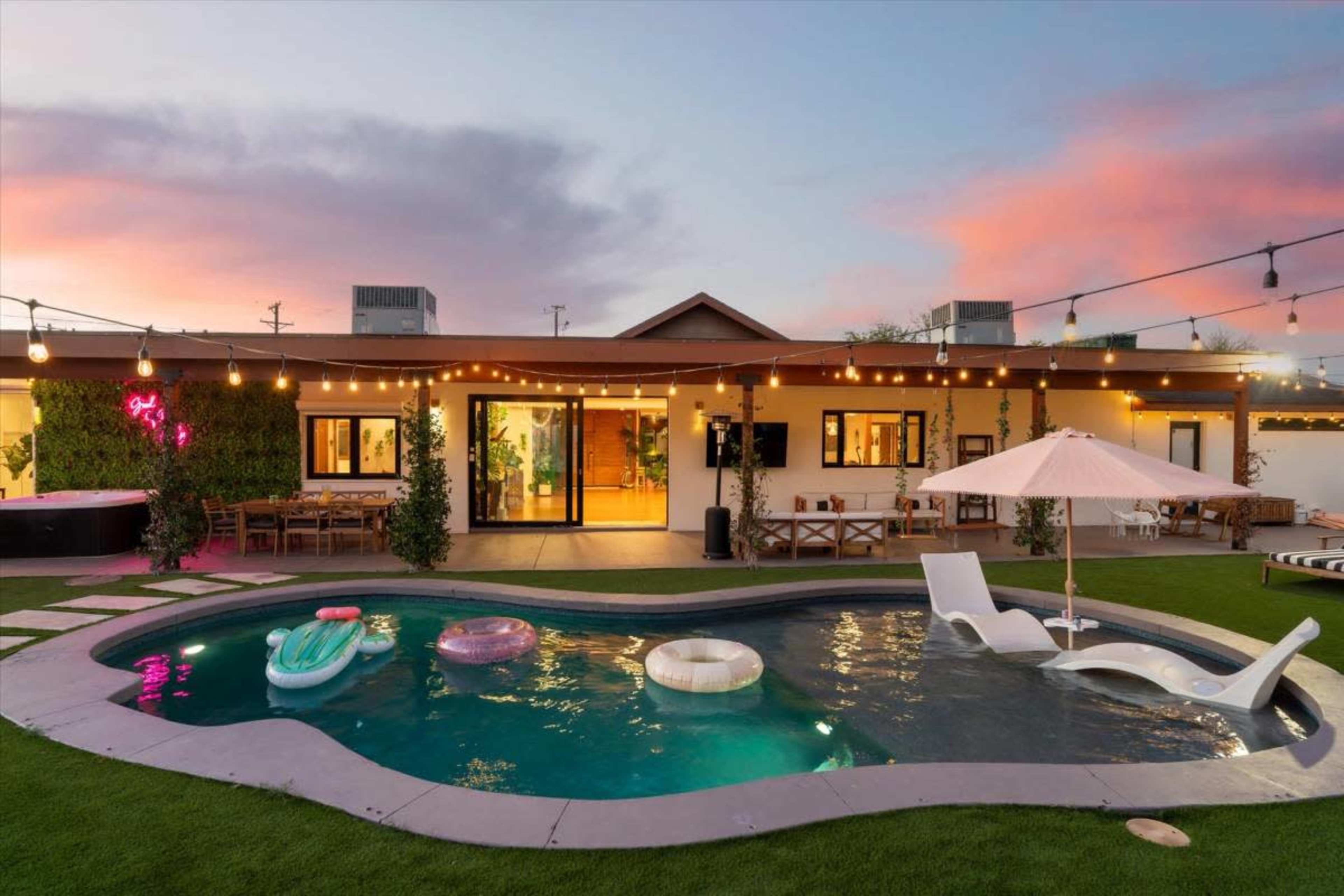 The image showcases a backyard with a swimming pool surrounded by lounge chairs and inflatable pool floats, adjacent to a modern house illuminated by string lights.