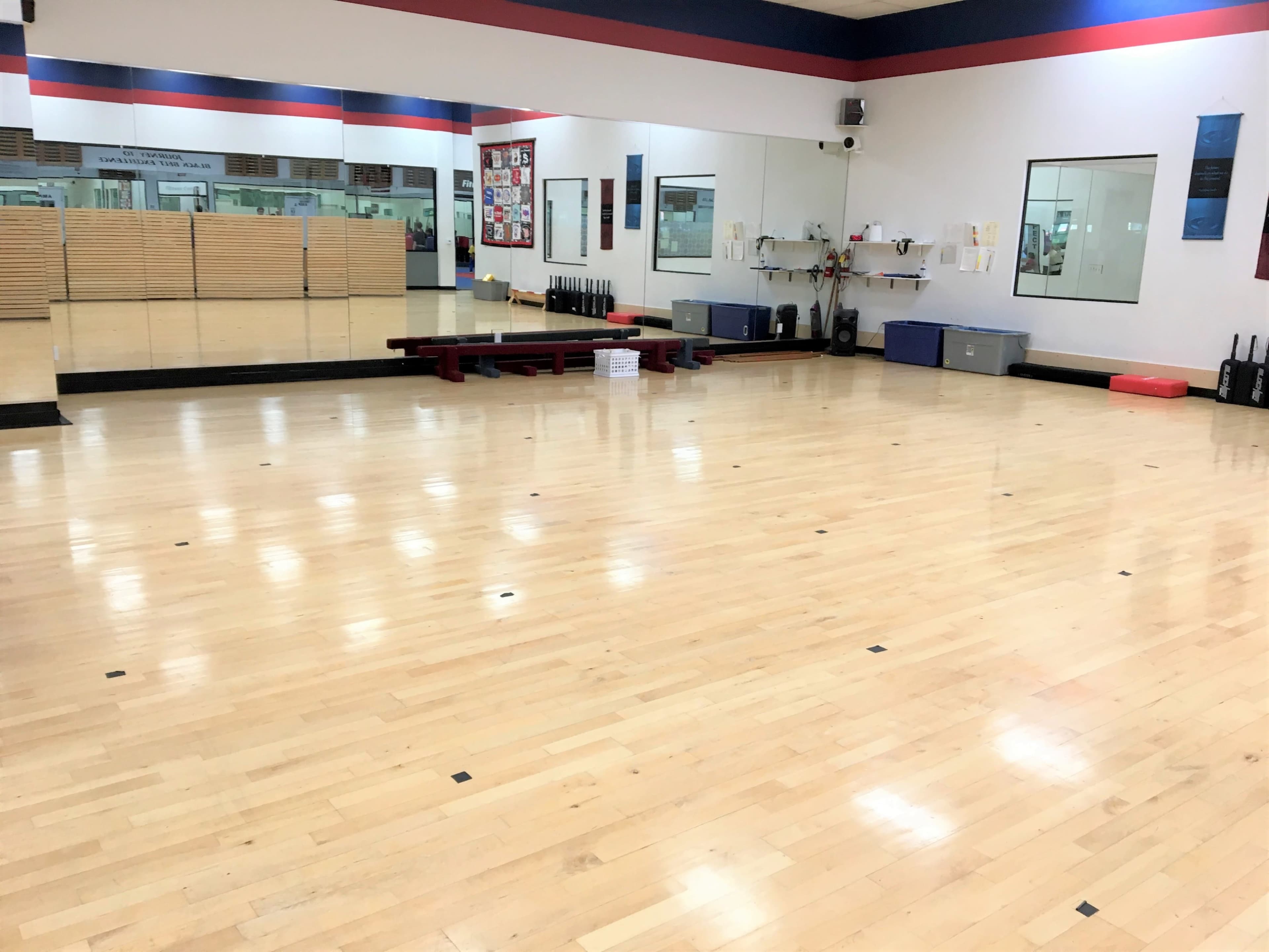 A spacious, empty gym studio with polished wooden flooring, mirrors on the wall, and exercise equipment neatly arranged in the background.
