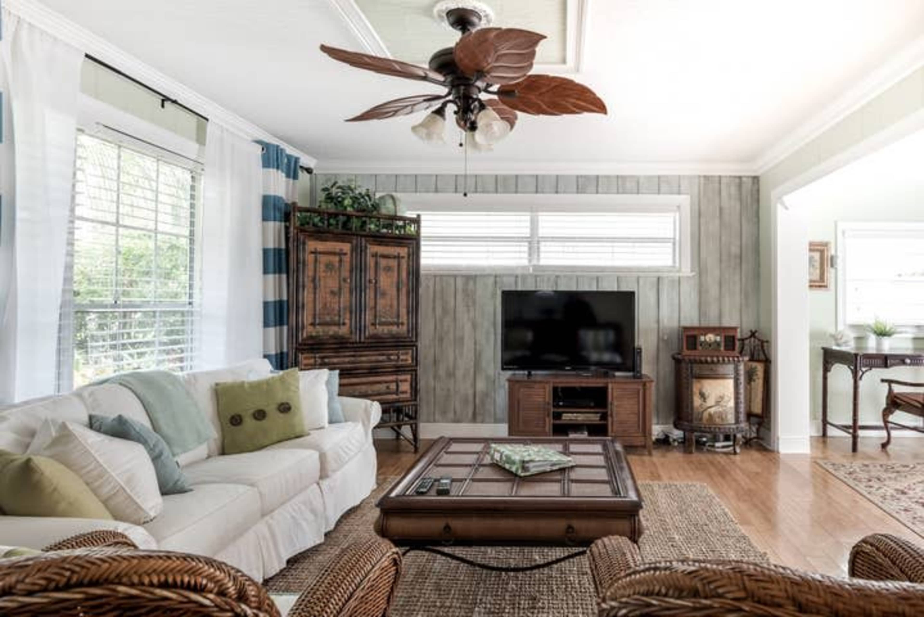 A living room featuring a white sofa with decorative pillows, a wooden coffee table, a television against a paneled wall, and wicker furniture.