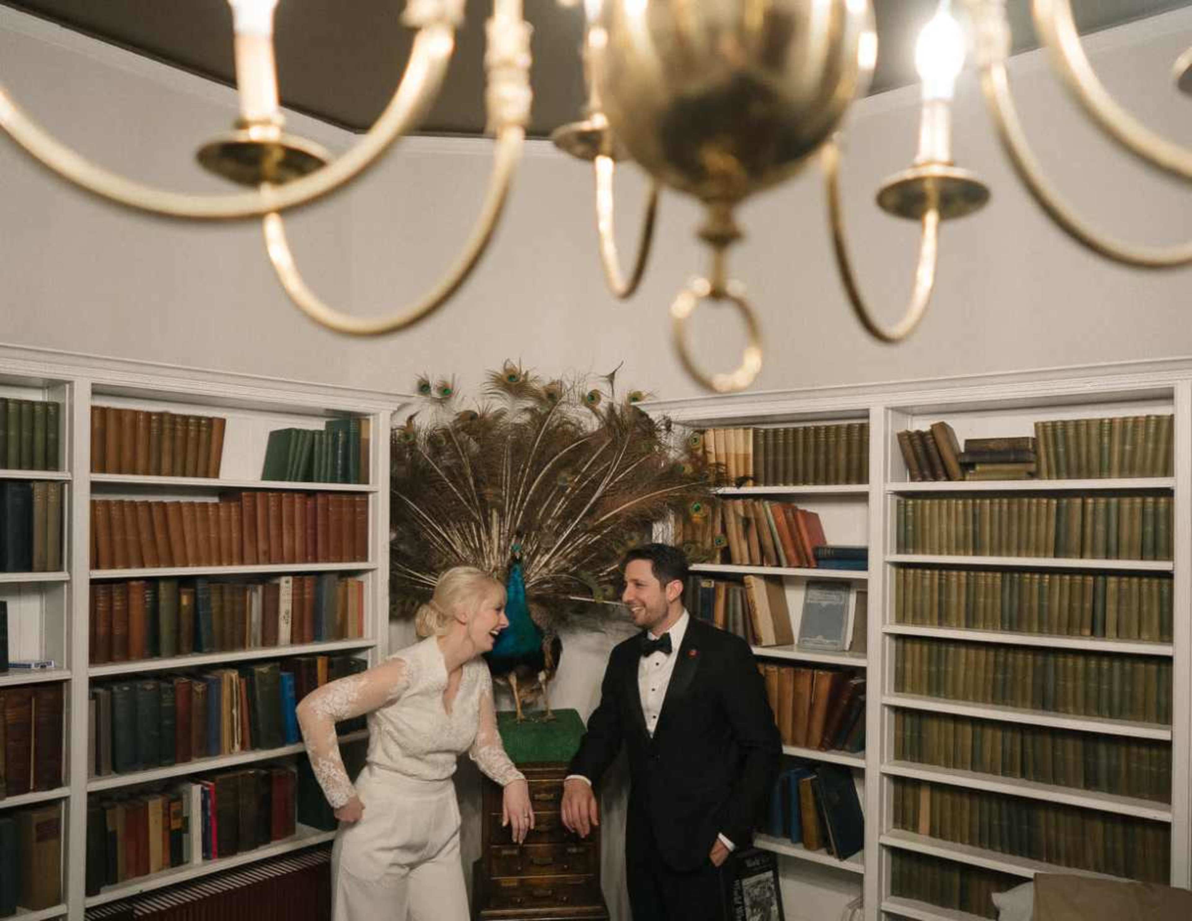 A couple stands in a library with bookshelves filled with various colored books, while a peacock is displayed prominently in the background.