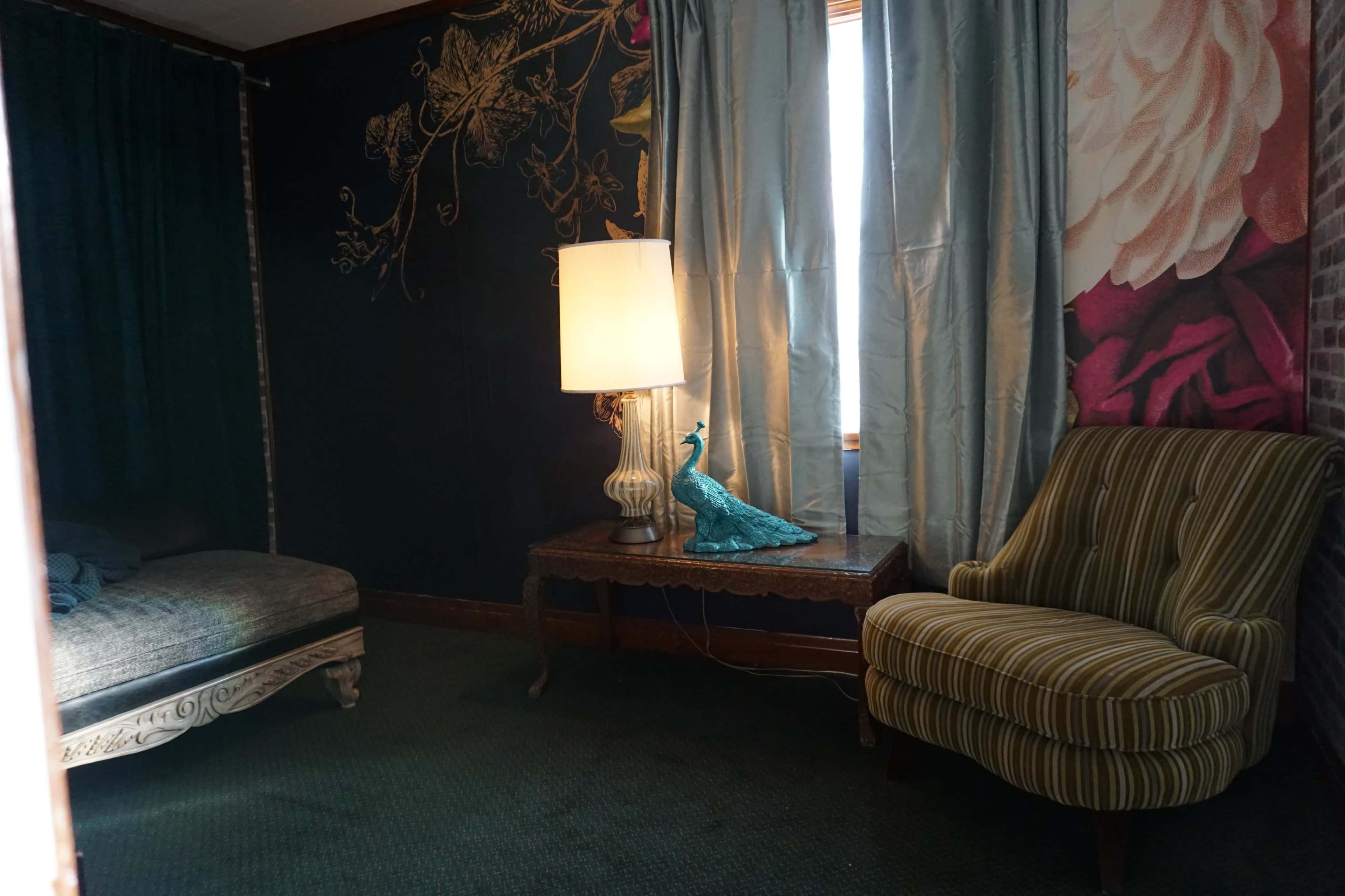 A corner of a room featuring a striped armchair, a patterned lamp on a side table, and a peacock figure, with floral wallpaper and curtains in the background.