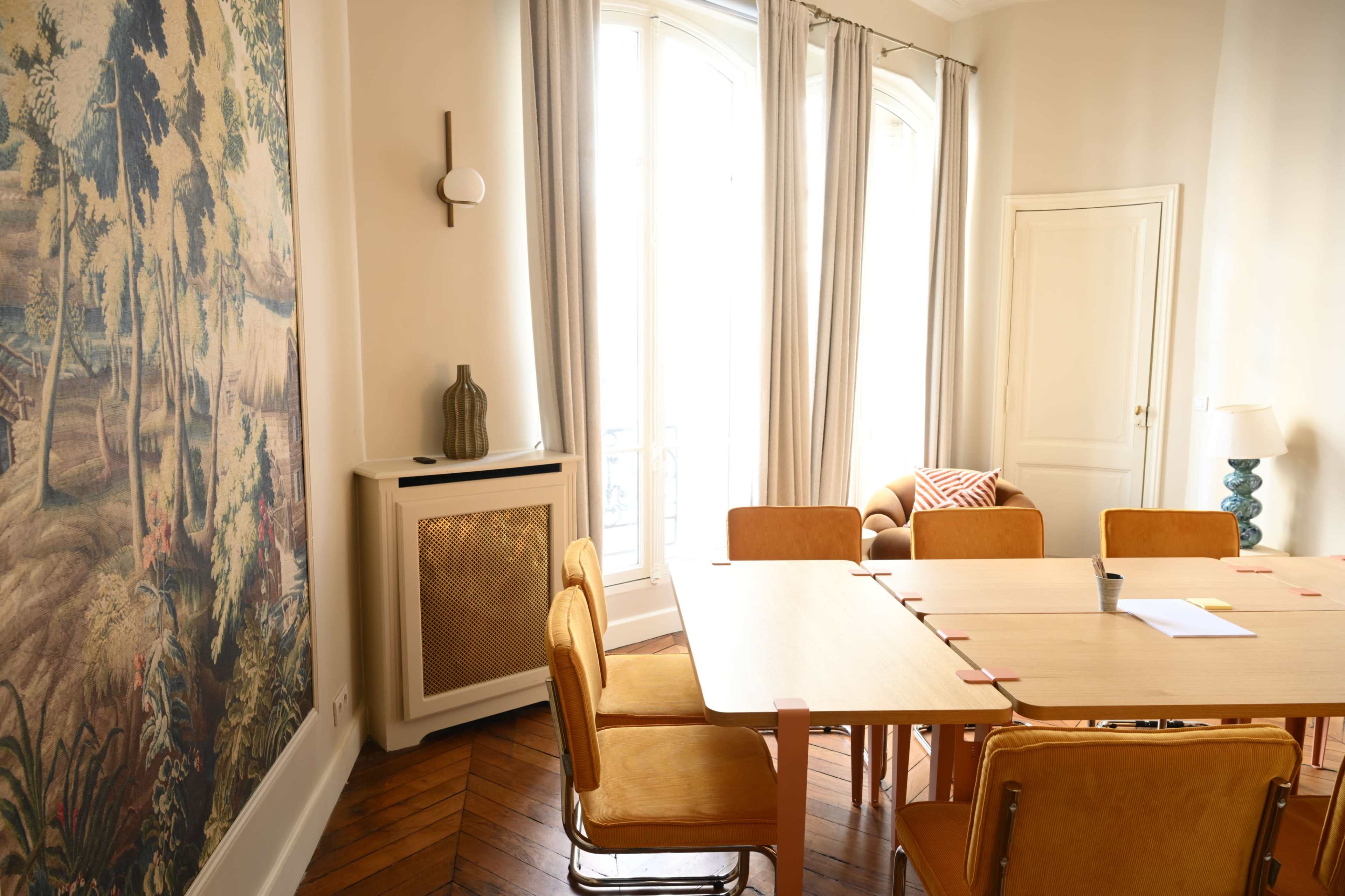 The image shows a brightly lit dining area with a wooden table surrounded by tan chairs, a large tapestry on the wall, and large windows that allow natural light to enter.
