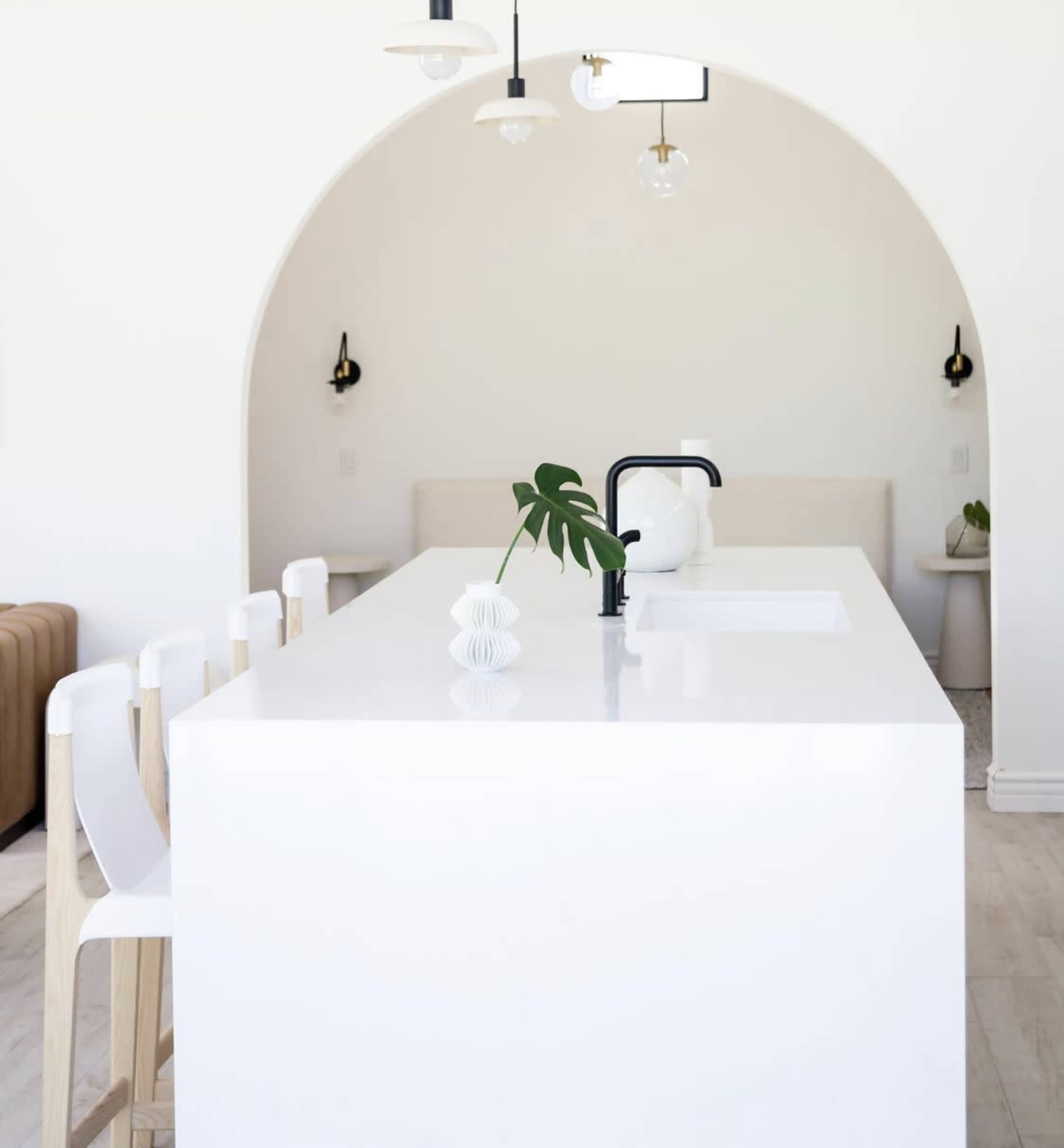 A minimalist kitchen with a large white island, black faucet, and light fixtures, set against an archway leading to a seating area.