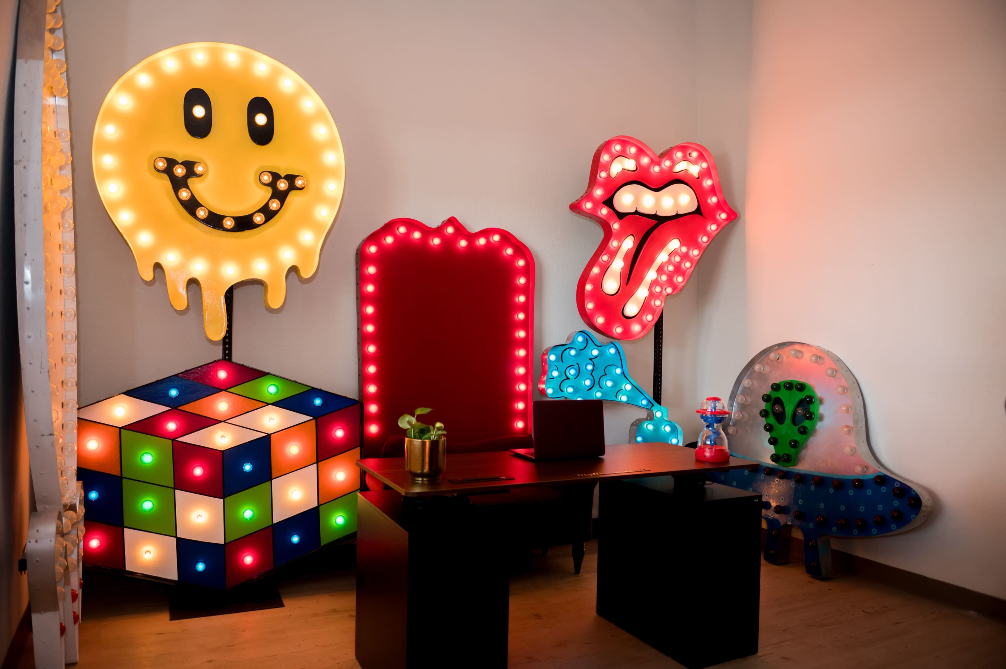 The image features a corner of a room decorated with colorful, illuminated signs, including a smiling face, a tongue-out mouth, a Rubik's Cube, and a ghost-like figure, alongside a dark desk.