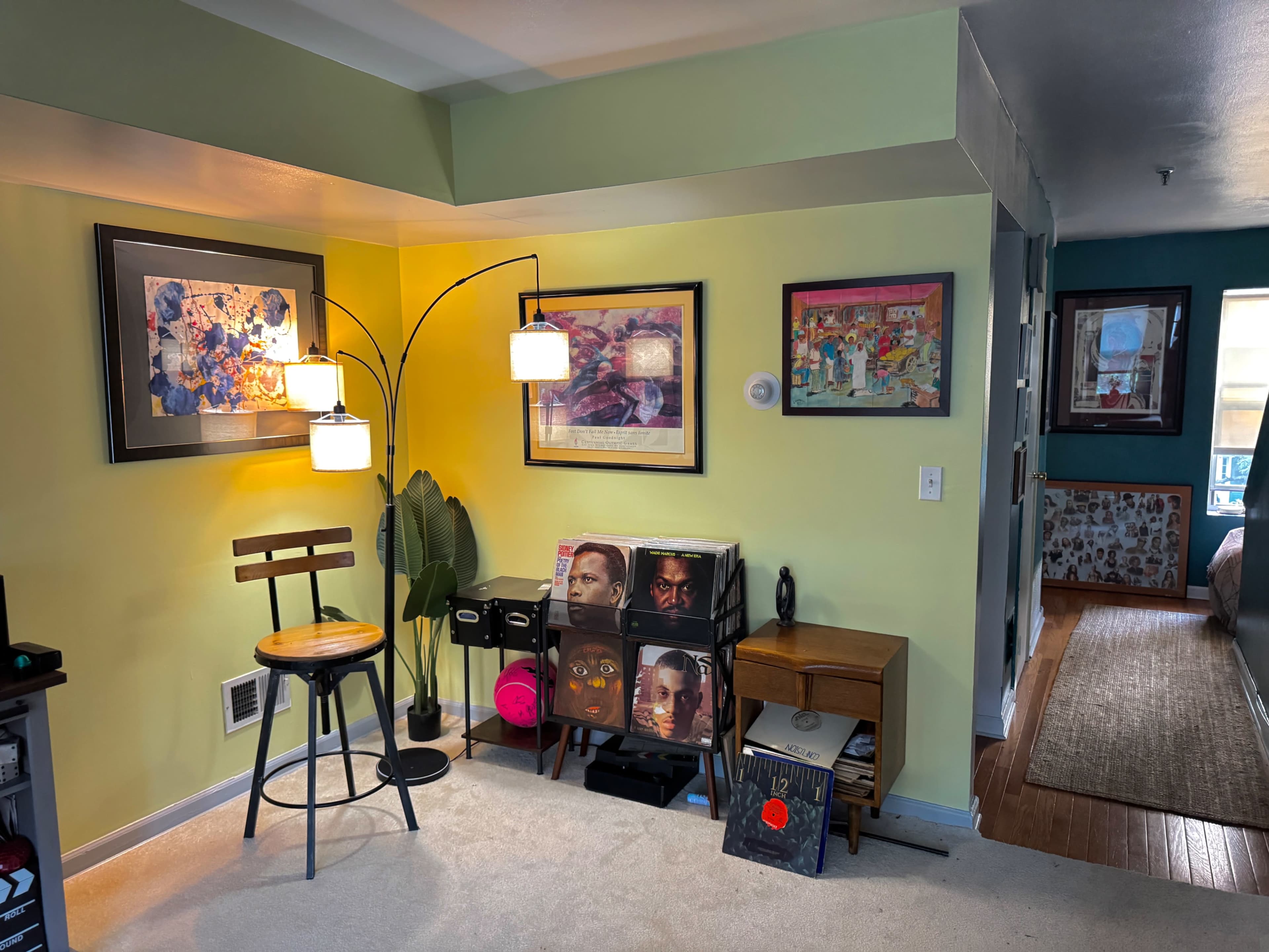 The scene shows a colorful living space featuring a small sitting area with a lamp, a barstool, framed artwork on the walls, and a display of vinyl records and decorative items.