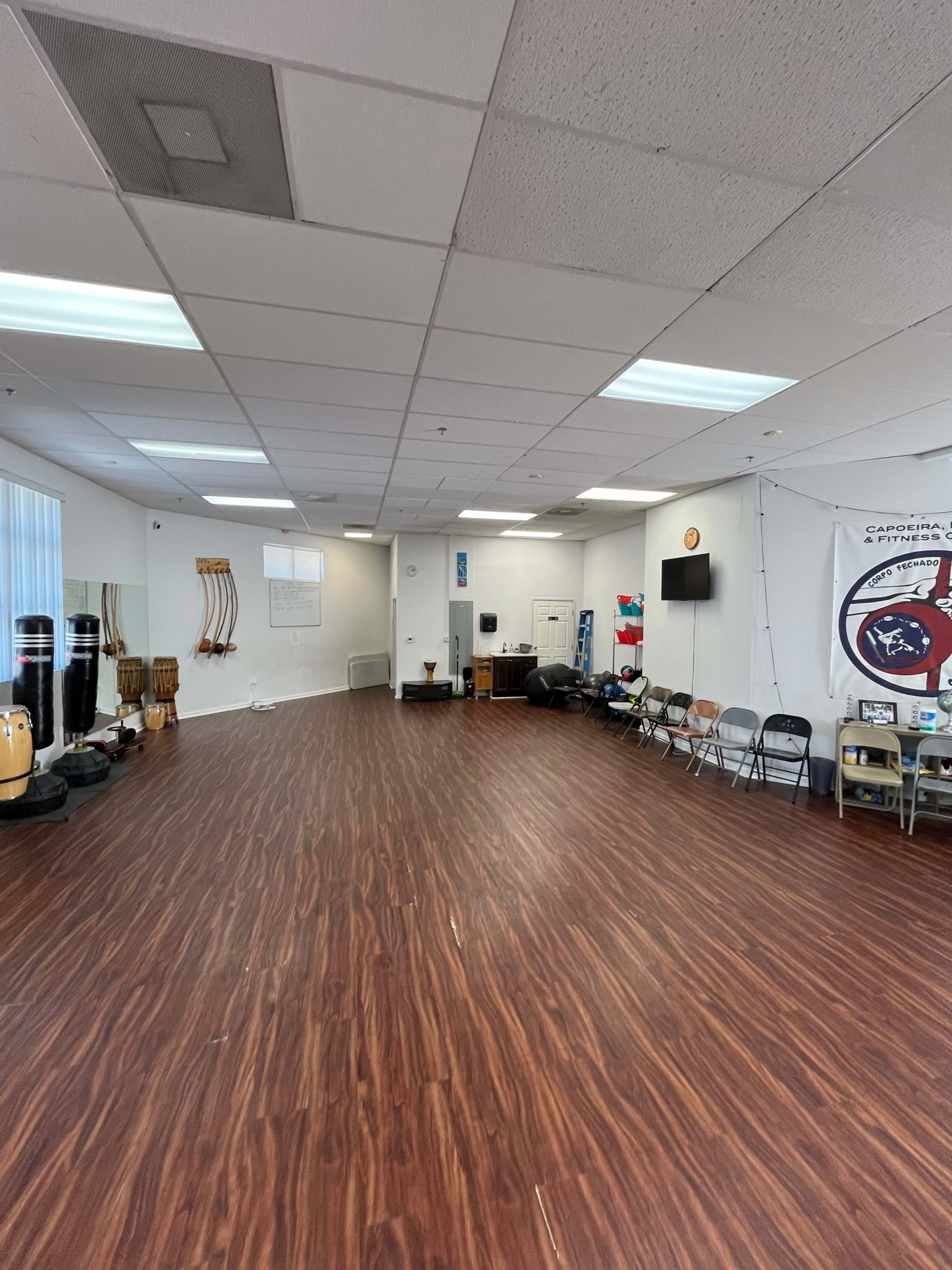 The image shows a spacious, empty room with wooden flooring, featuring musical instruments and chairs arranged along the sides.