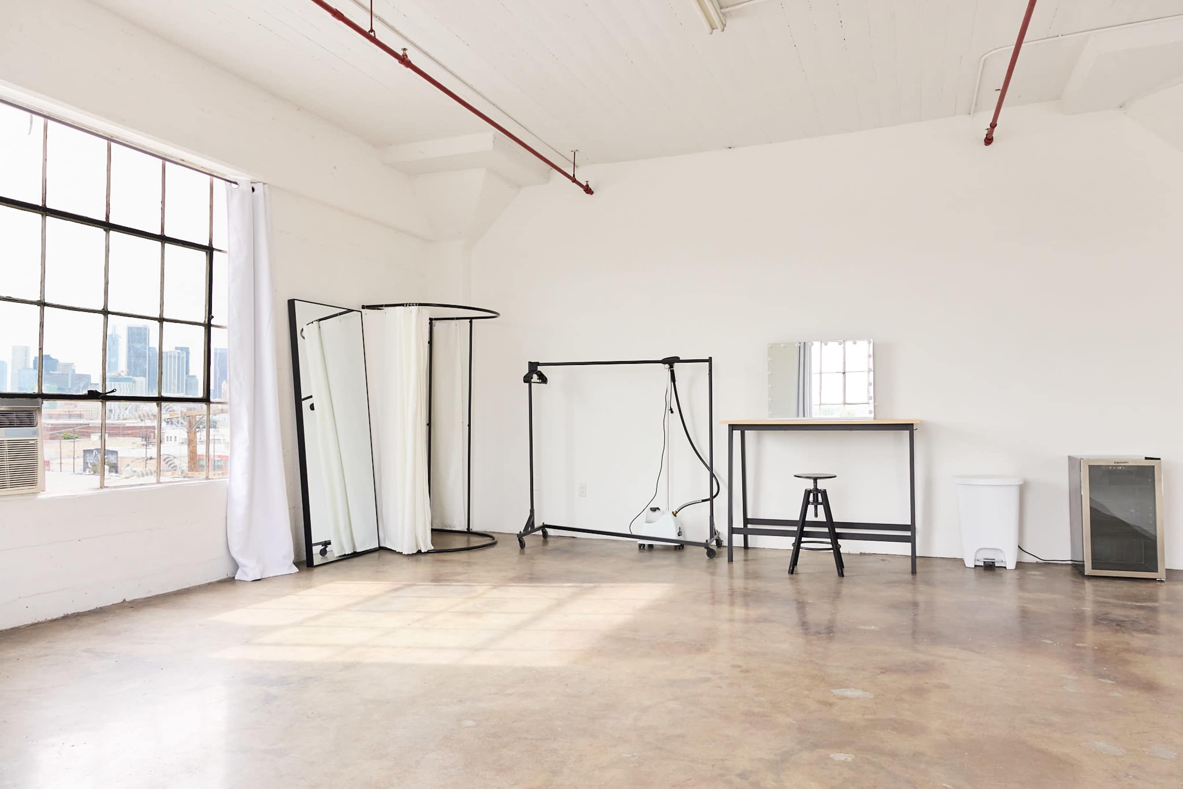 The image shows a spacious, empty room with large windows, a few mirrors, a standing light, a stool, a table, and a small refrigerator.