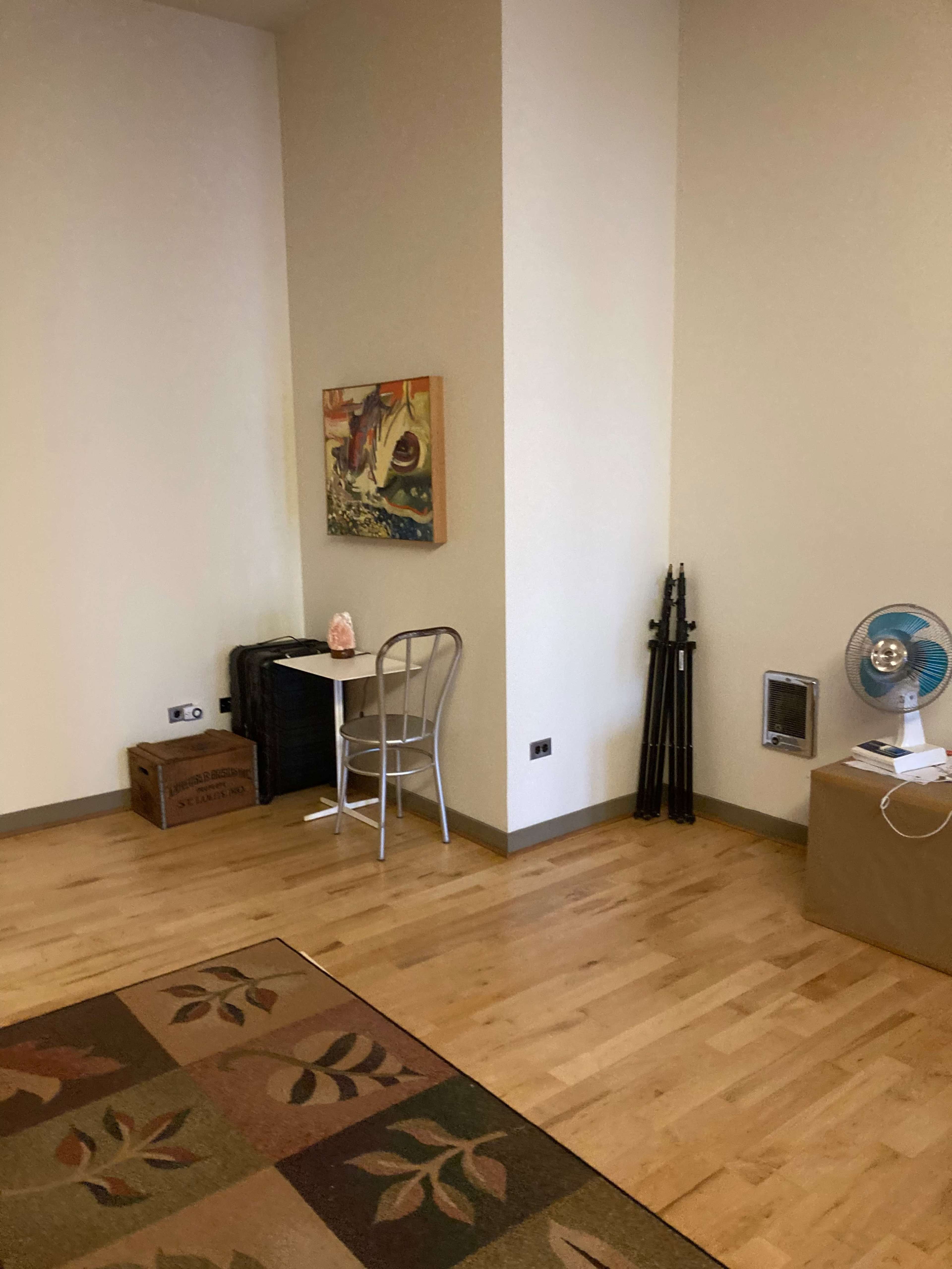 The image shows a corner of a room with a wooden floor, a small table and chair, an art piece on the wall, a fan on the floor, and some storage boxes nearby.