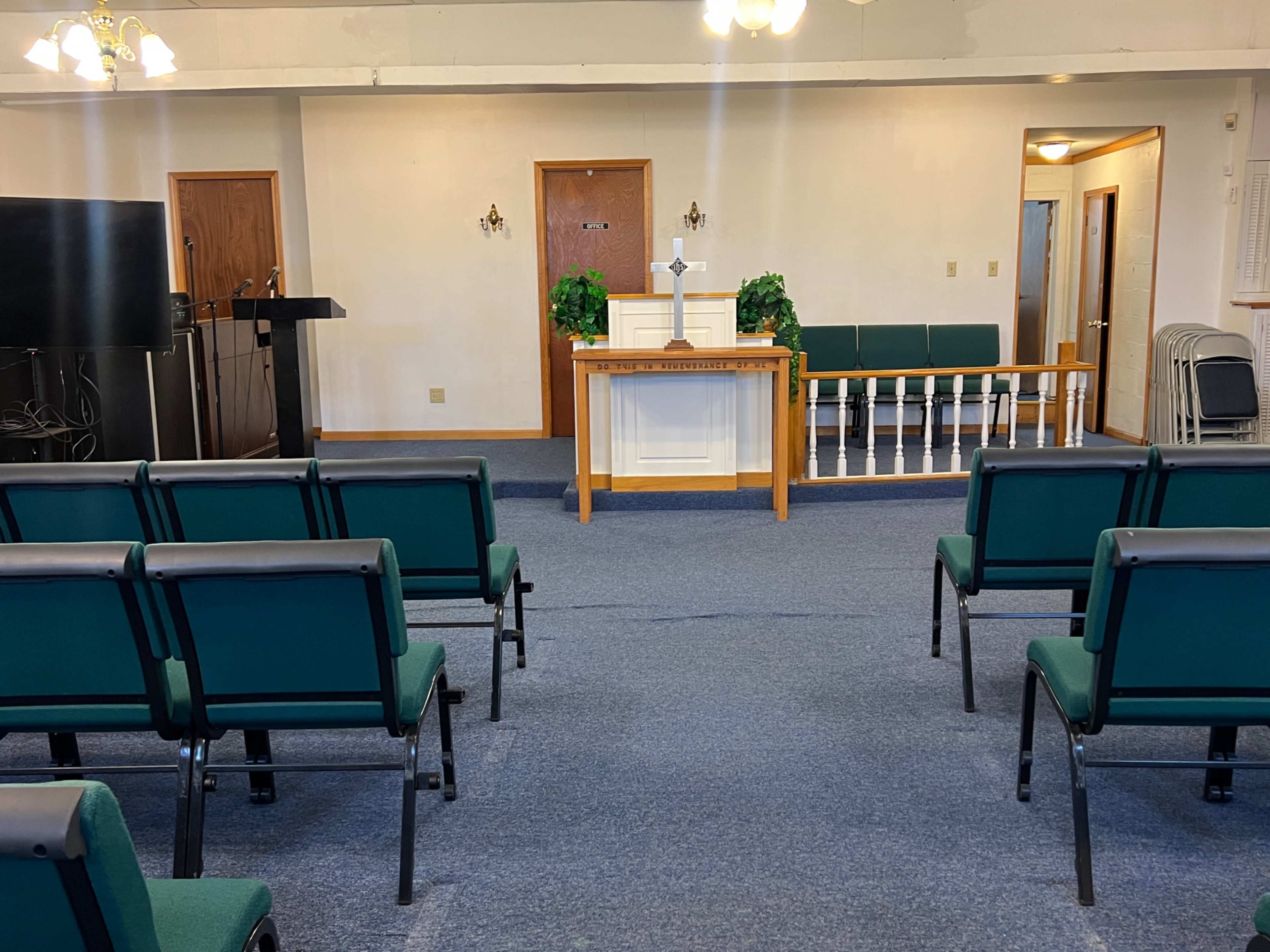 The image shows the interior of a small church with green chairs facing a wooden altar at the front.