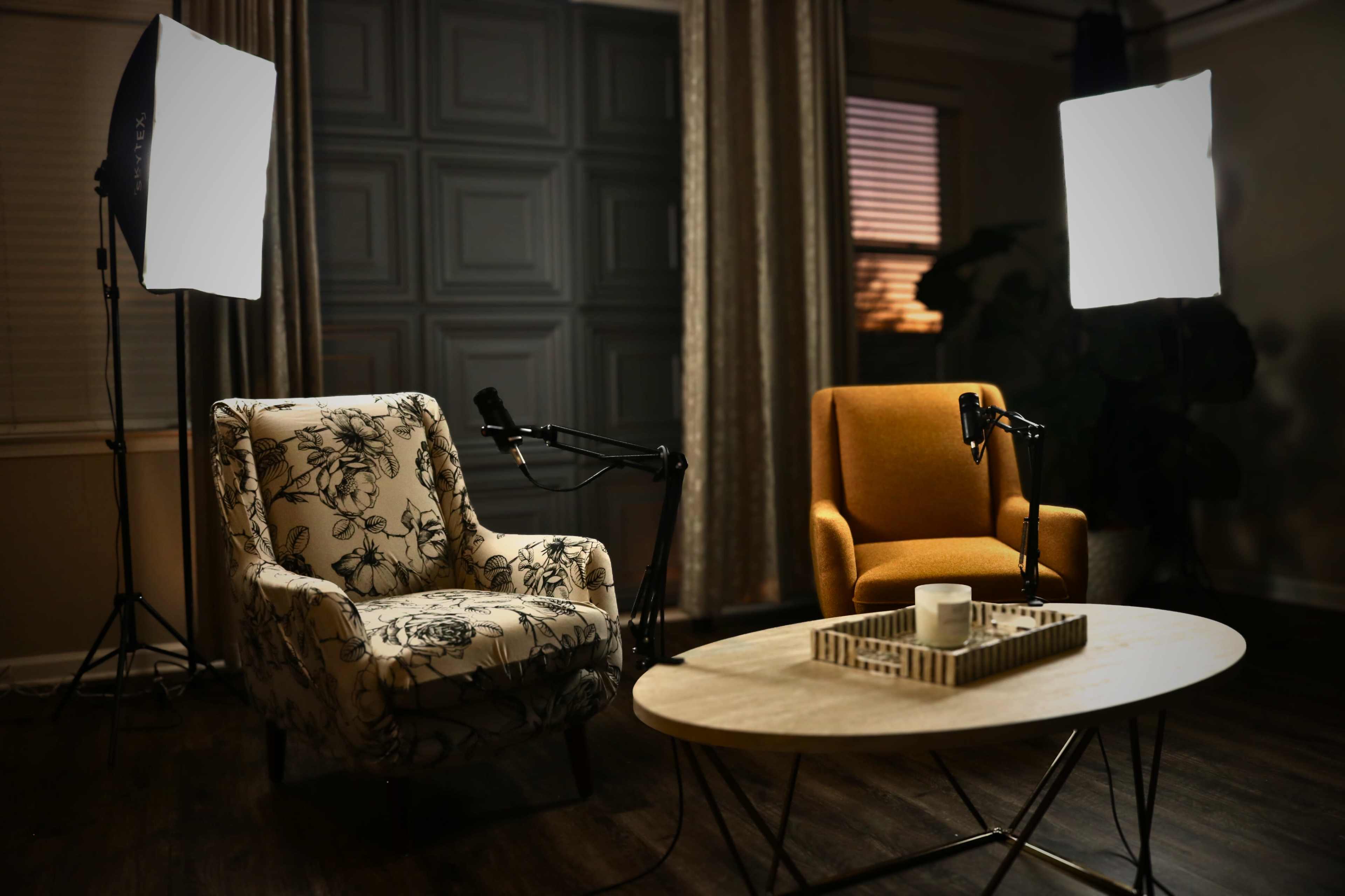 The image shows a small, well-lit interview setup featuring two armchairs, a round coffee table, and two microphones positioned in front of the chairs.