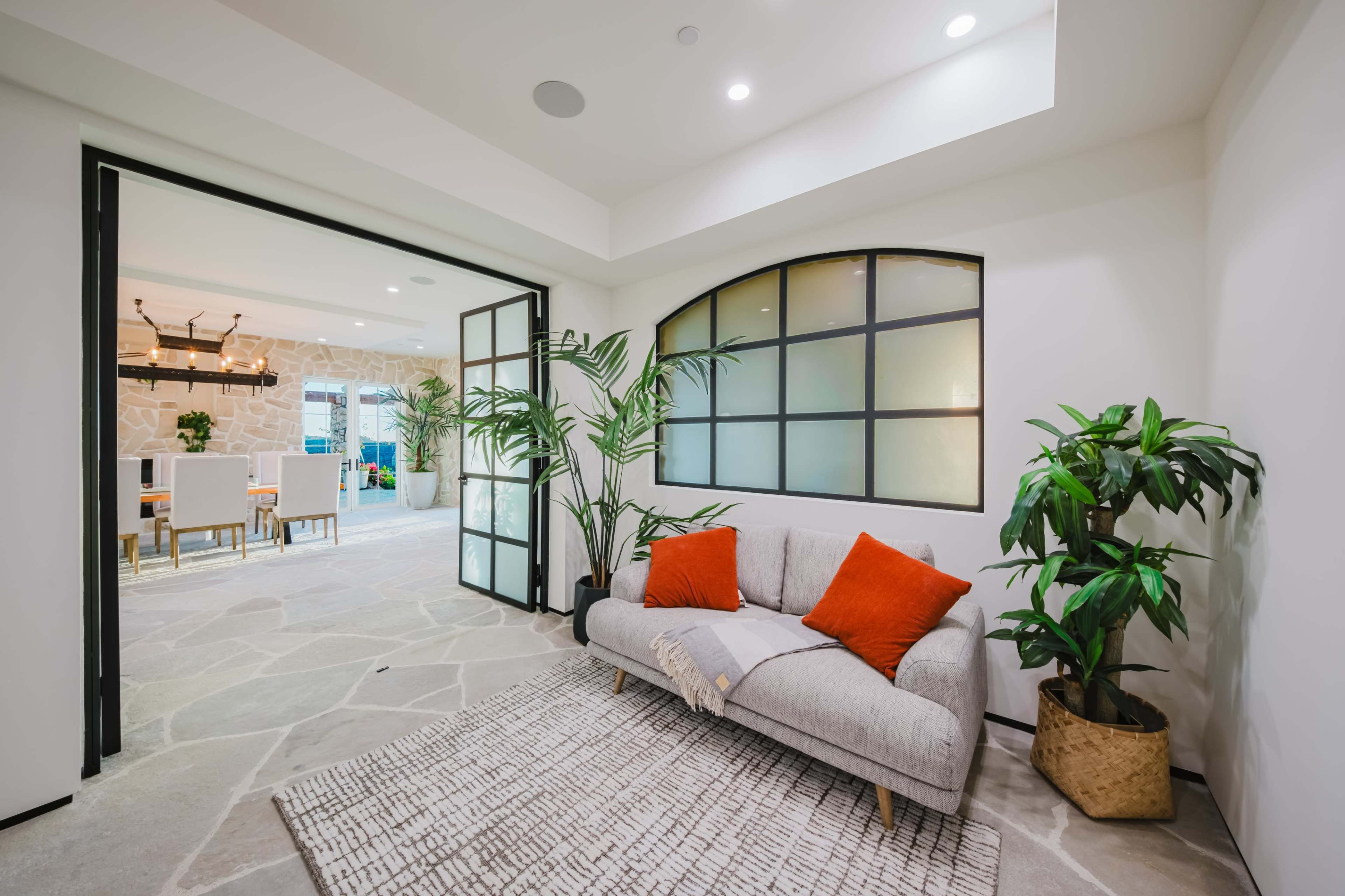 A minimalist living space features a light gray sofa with orange pillows, surrounded by indoor plants and an open doorway leading to another room.