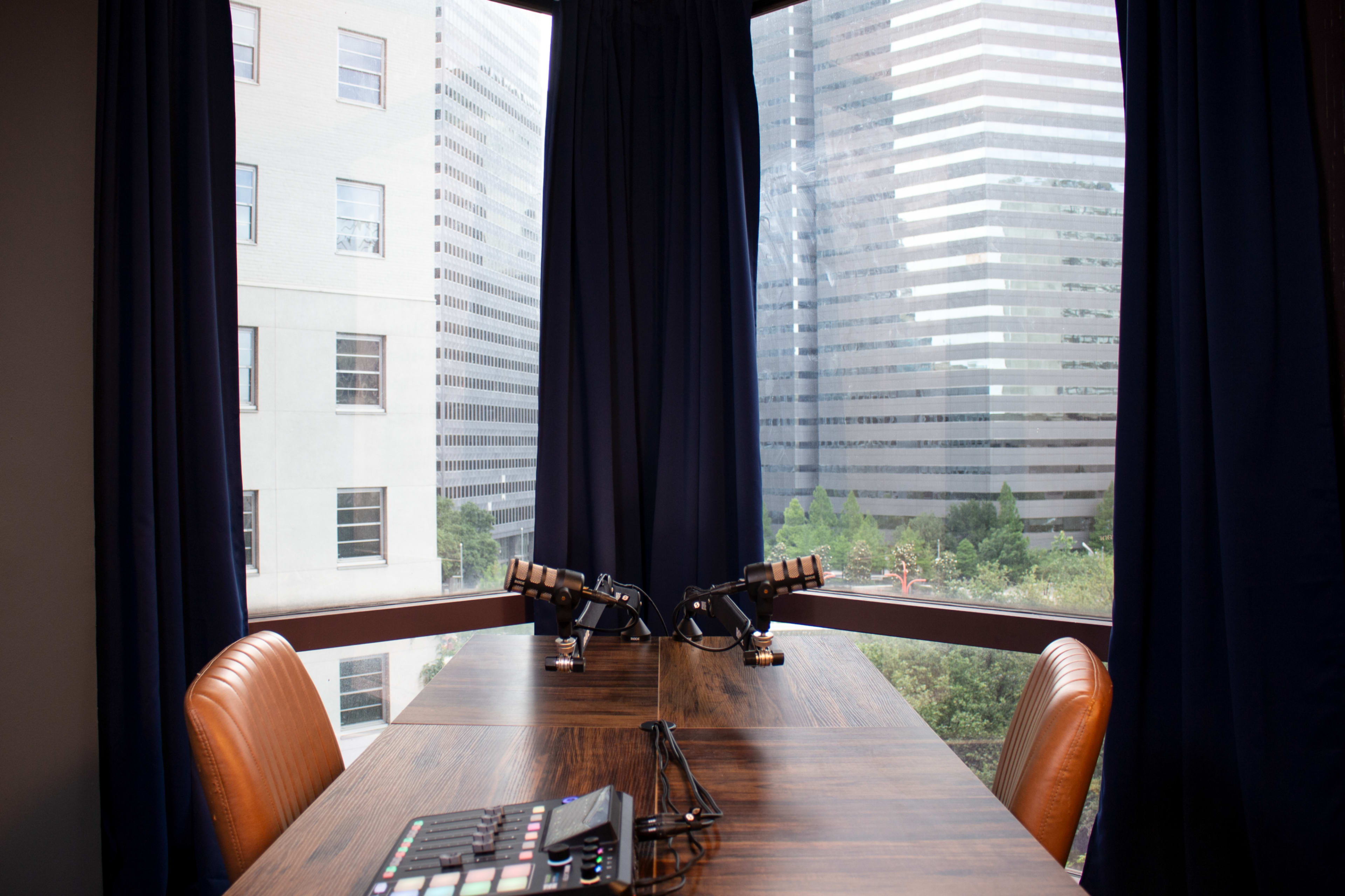 The image shows a modern conference room with a wooden table, two microphones, and large windows overlooking tall buildings.