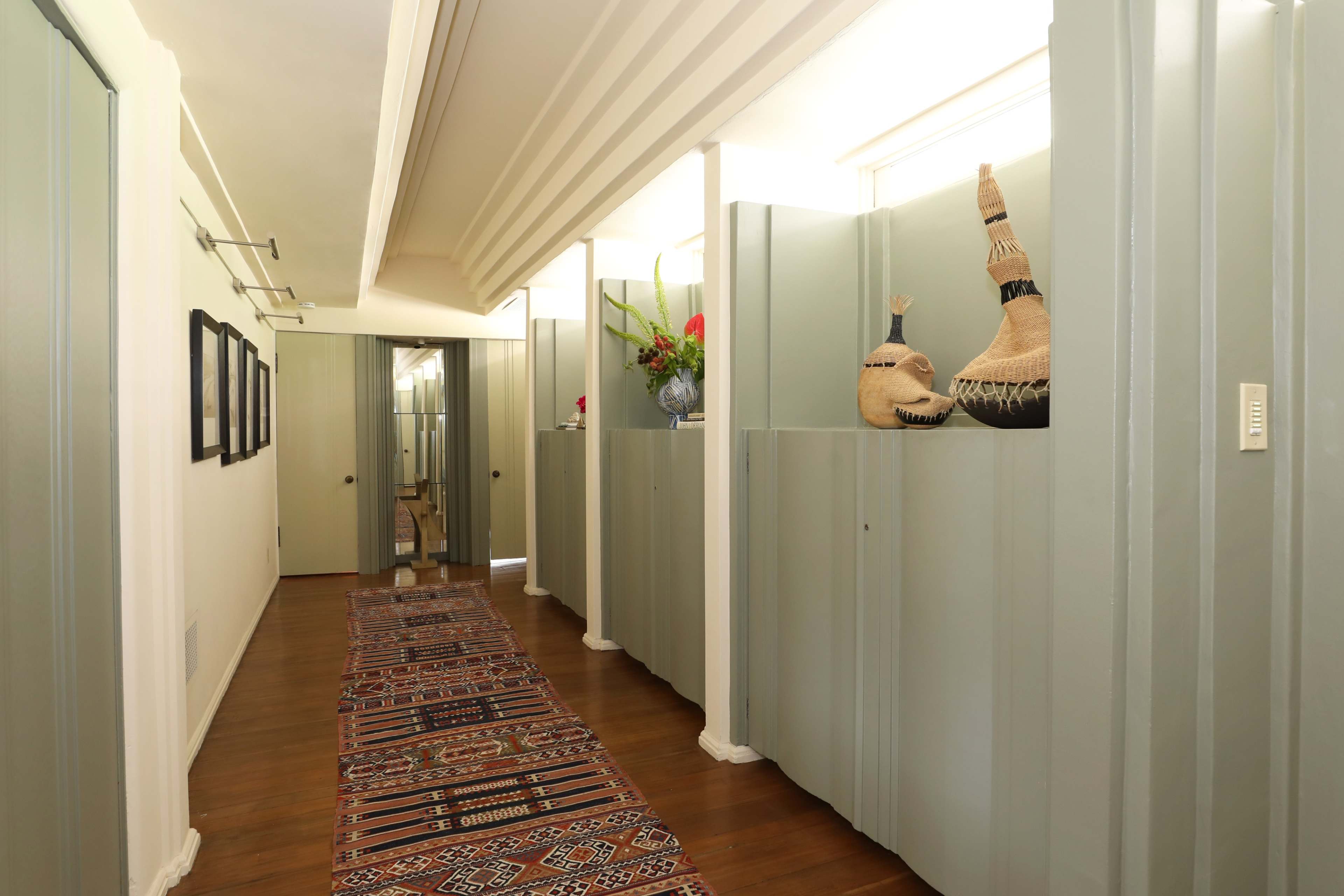 A narrow hallway features green partition walls, framed pictures, and decorative items on shelves, with a patterned rug running along the wooden floor.