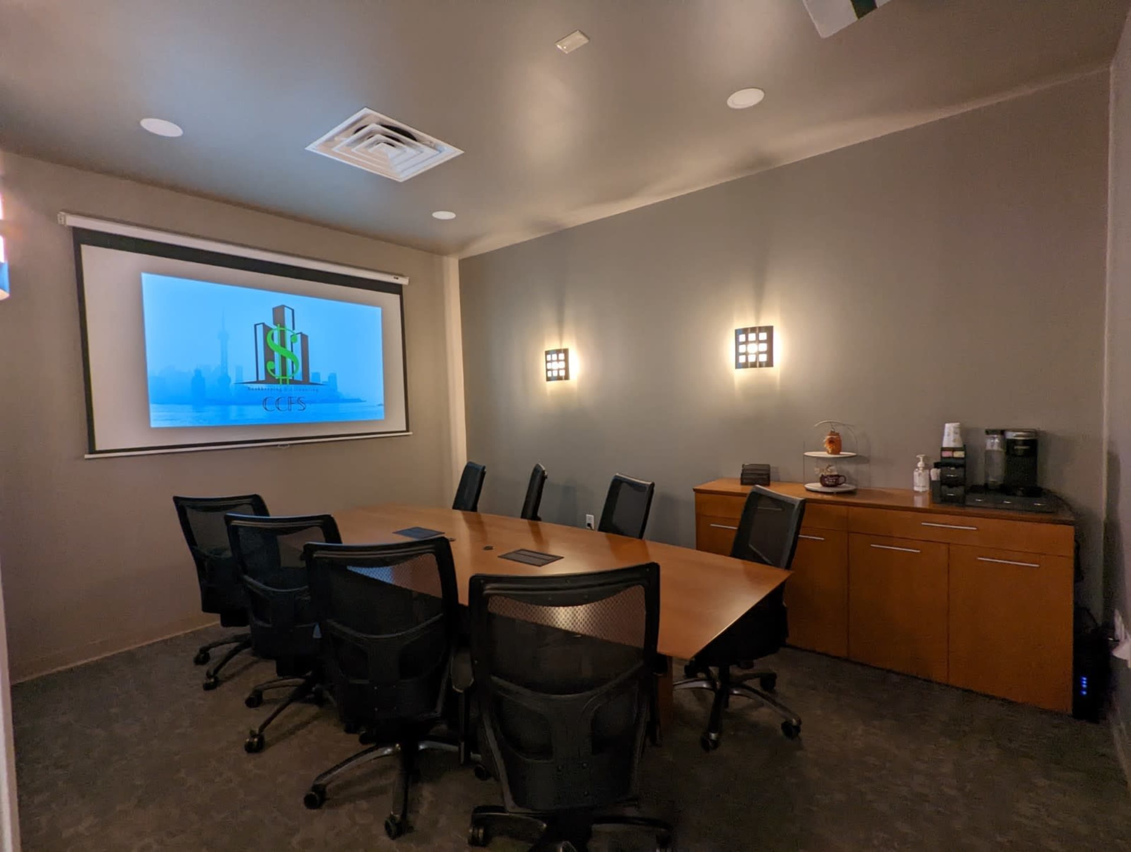 A conference room with a large screen displaying a logo, a wooden table surrounded by black office chairs, and a side cabinet with a coffee machine.