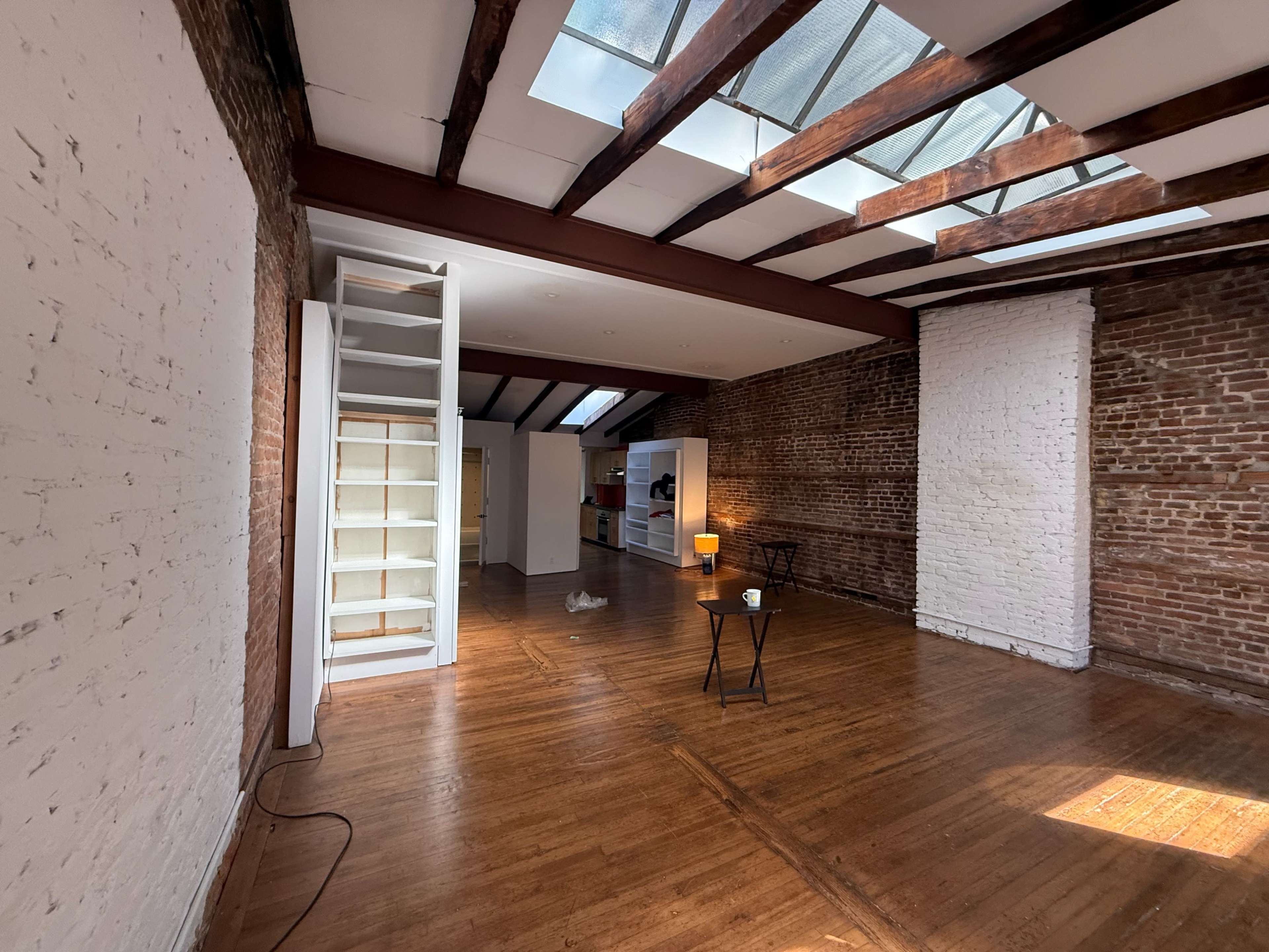 The image shows a spacious, empty loft featuring exposed brick walls, wooden beams, and large skylights that illuminate the wooden floor.