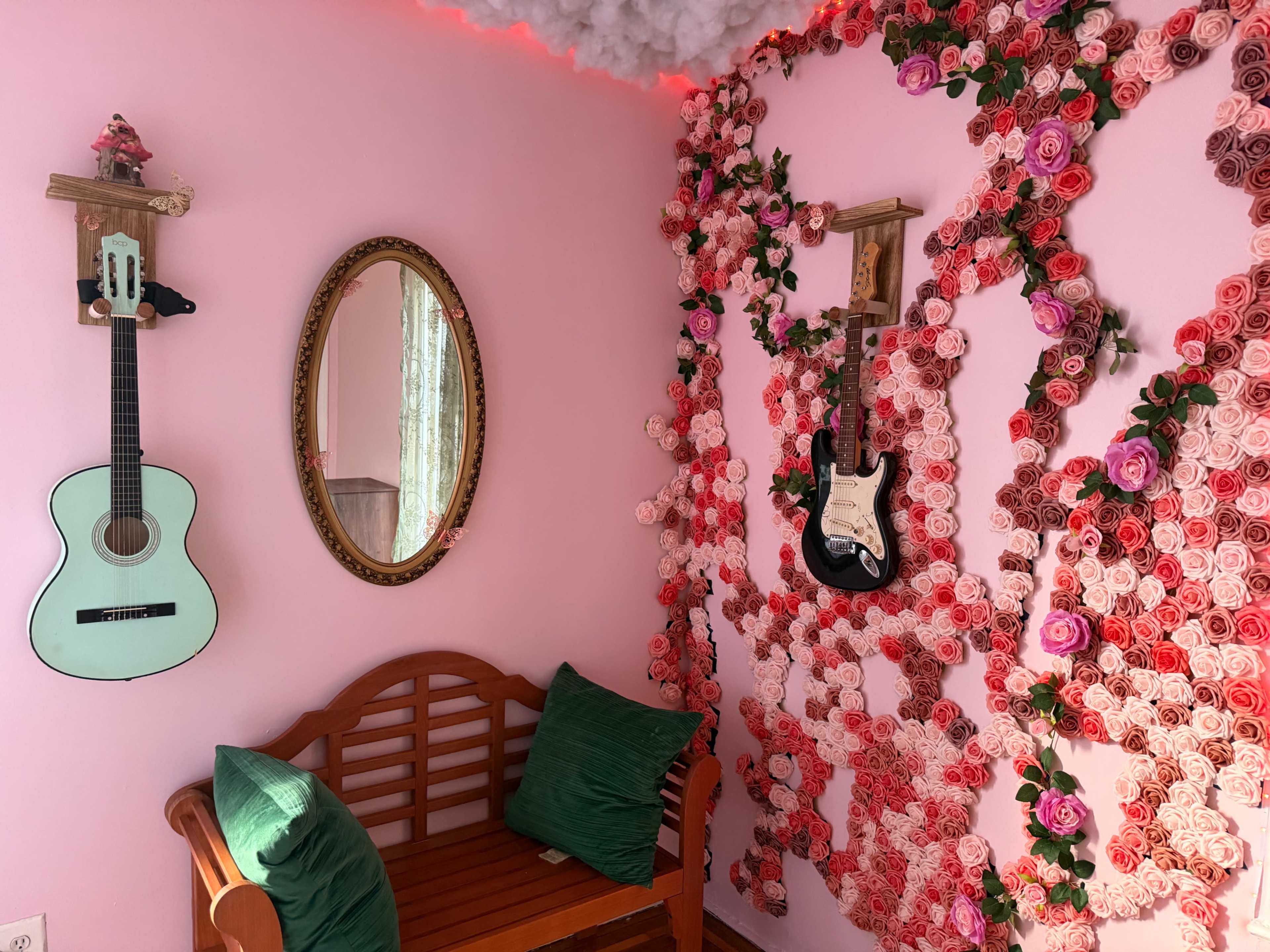 The image shows a cozy corner with a wooden bench, a circular mirror, and two guitars mounted on a pink wall adorned with artificial flowers.