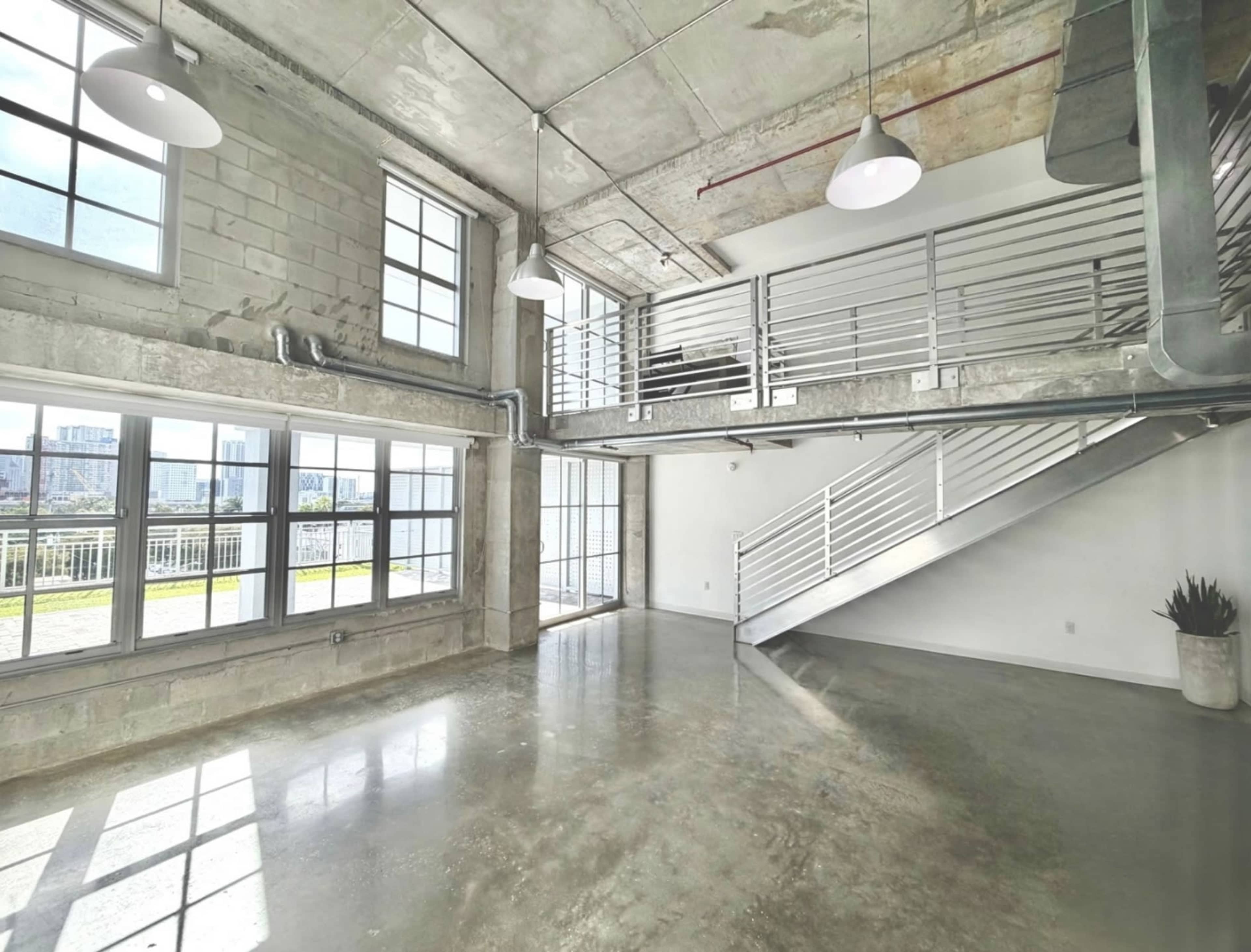 The image shows an industrial-style interior space with large windows, concrete walls, and a staircase leading to a second level.