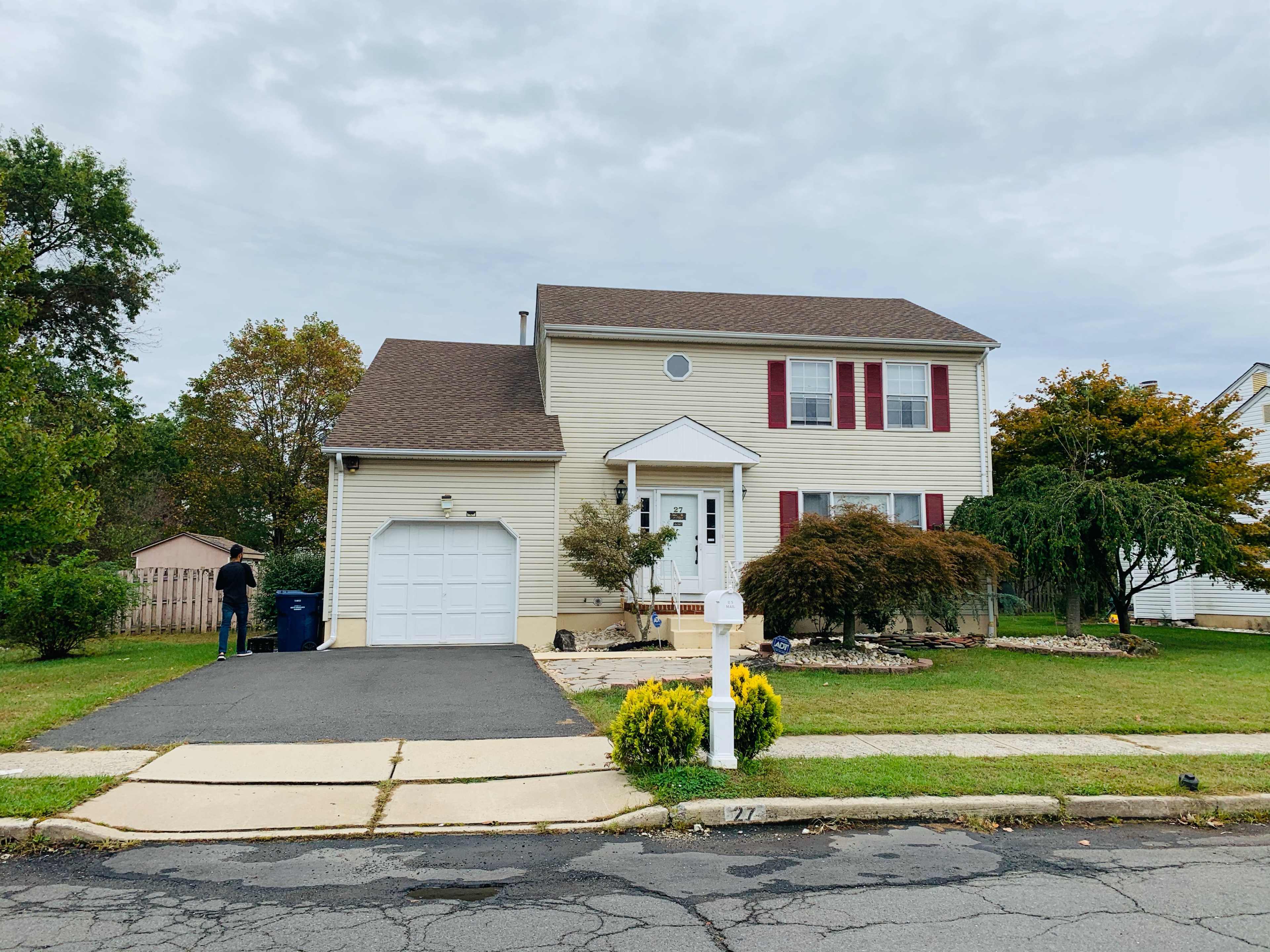 A two-story house with beige siding, red shutters, and a garage is situated on a neatly maintained lawn.