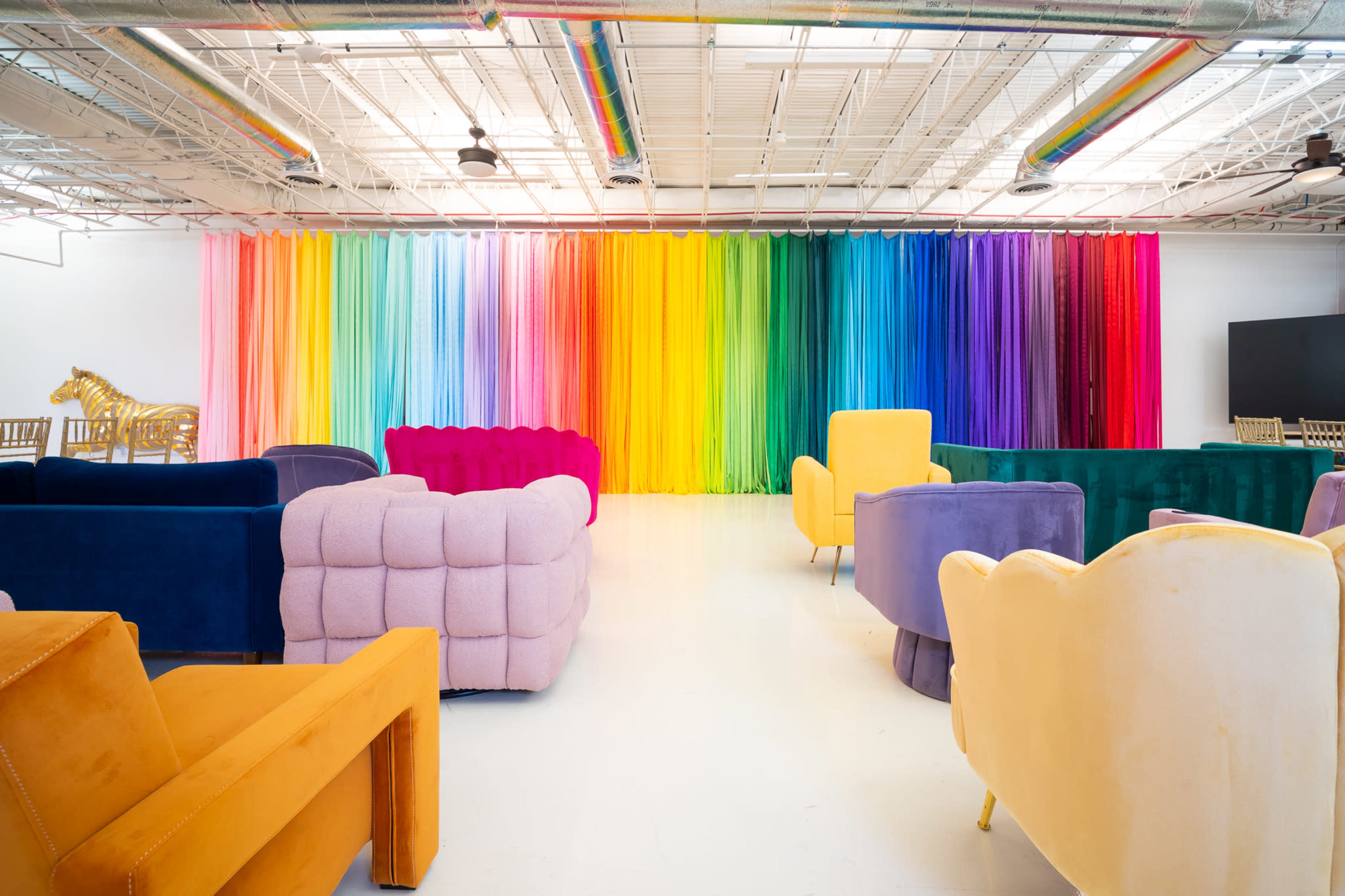The image shows a brightly colored room with a rainbow gradient backdrop and various plush seating arrangements in contrasting colors.