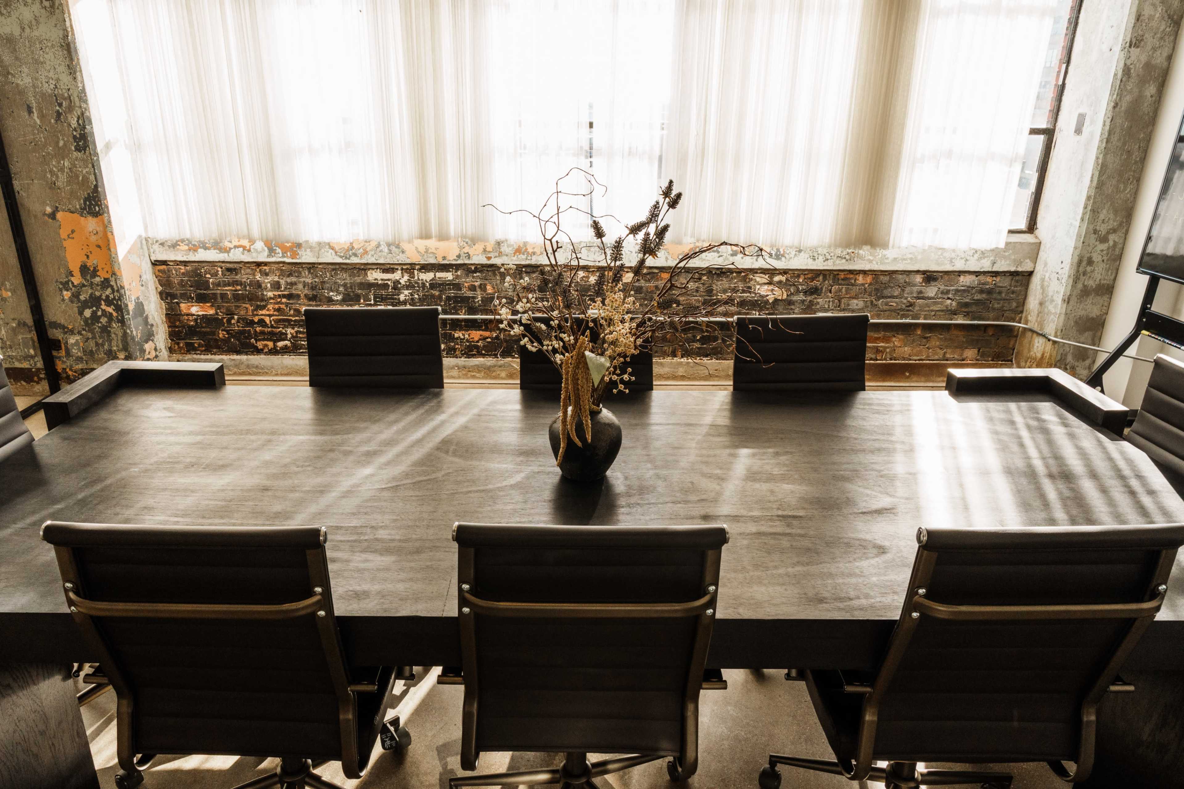 A dark wooden conference table is set in a bright room with large windows, surrounded by several black leather chairs and decorated with a vase of dry branches.