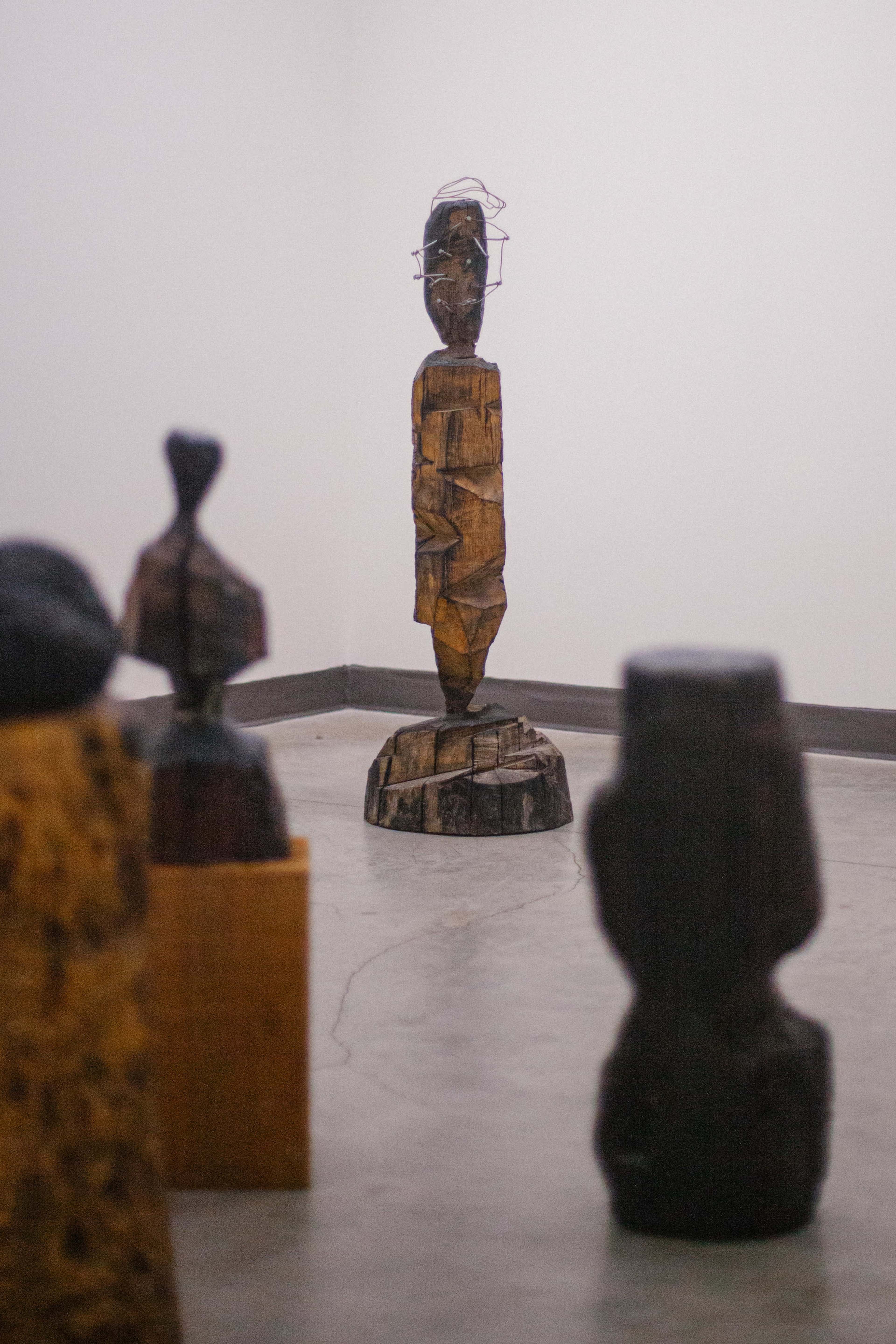 The image shows a wooden sculpture standing on a pedestal in a gallery, surrounded by other abstract wooden sculptures.