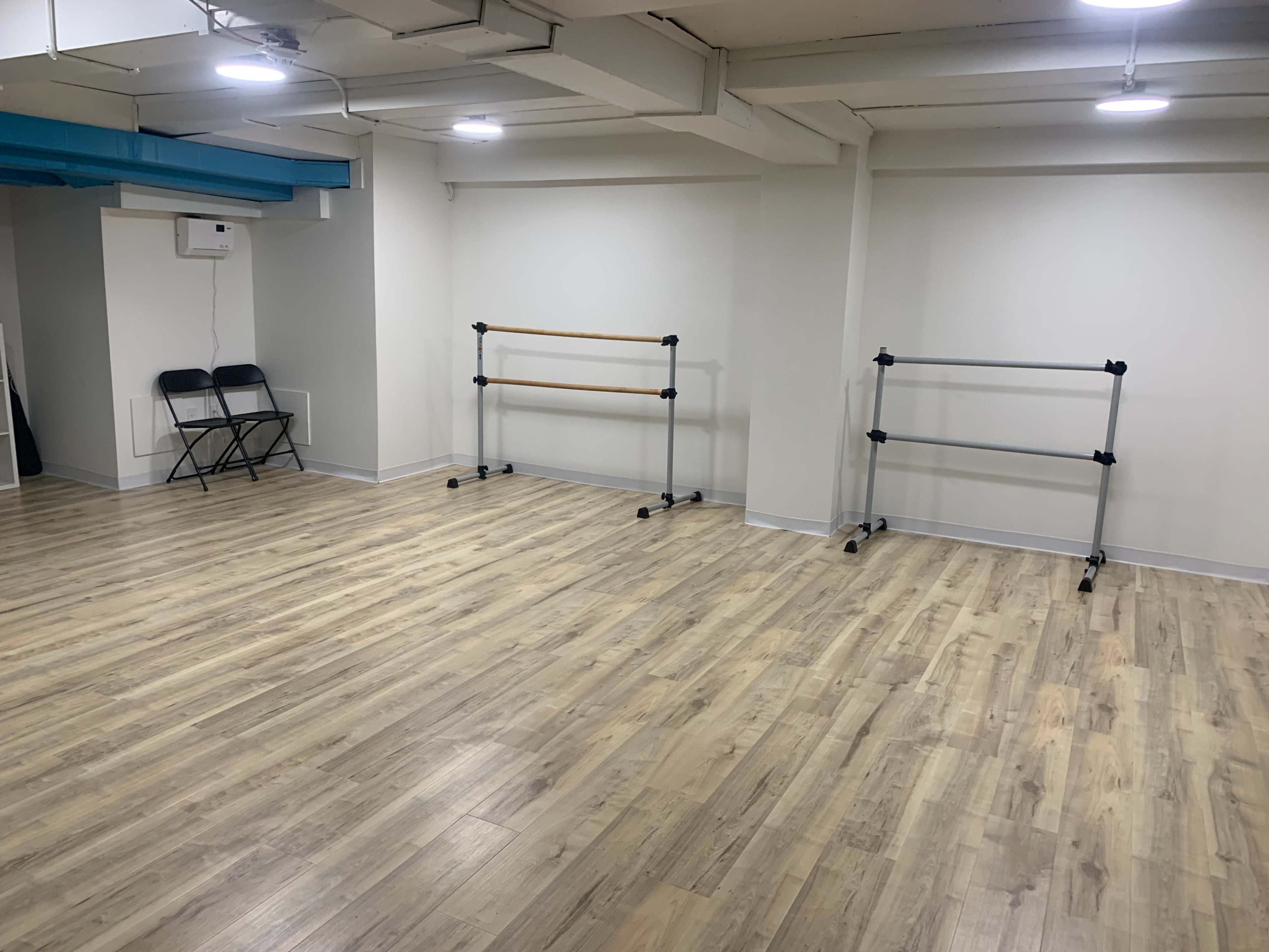 The image shows a spacious, empty room with wooden flooring, two ballet barres on rolling stands, and a couple of folding chairs against one wall.