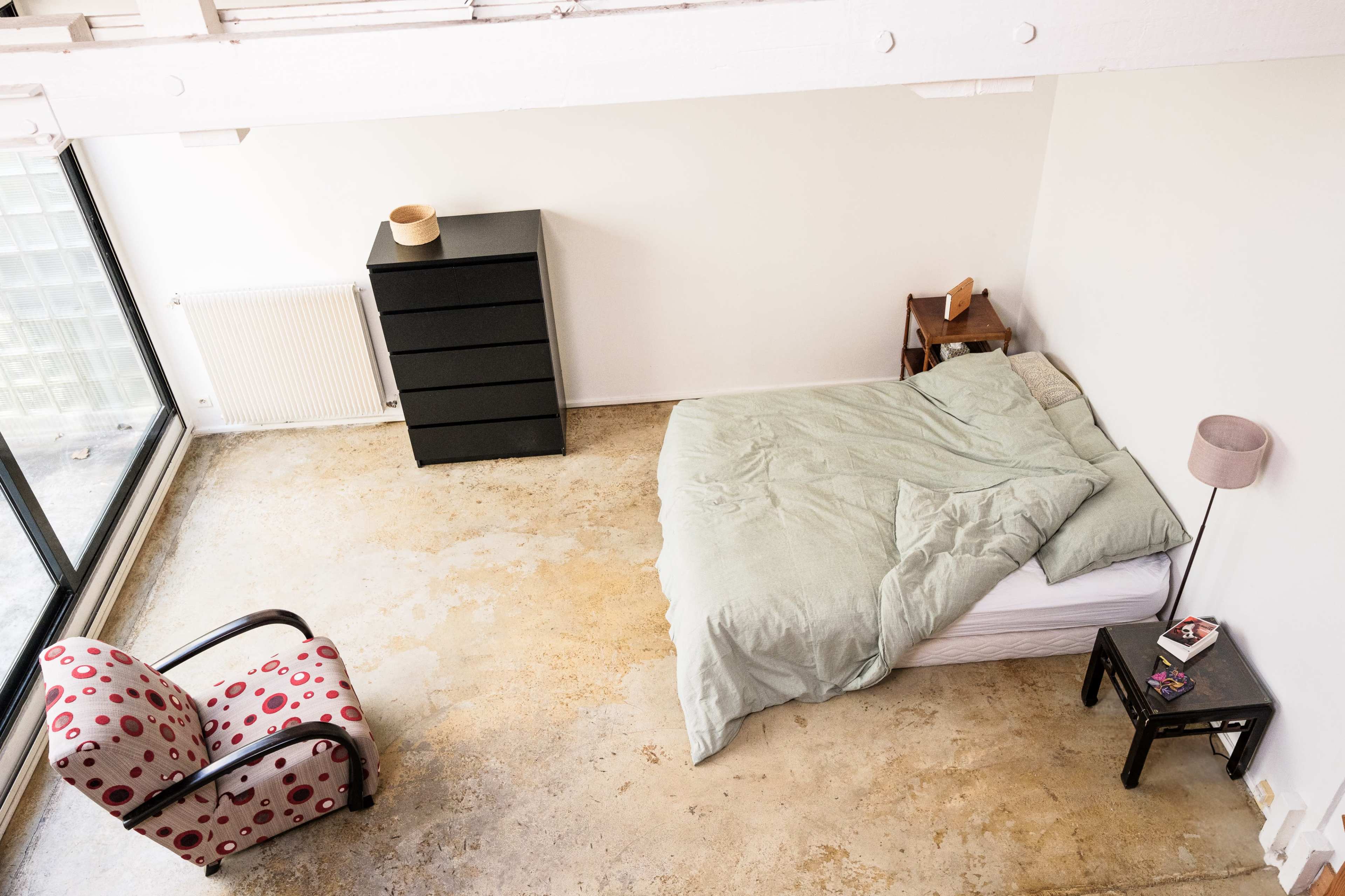 A minimalist bedroom features a bed with light green bedding, a black dresser, and a chair with a red and white pattern, set against a large window.