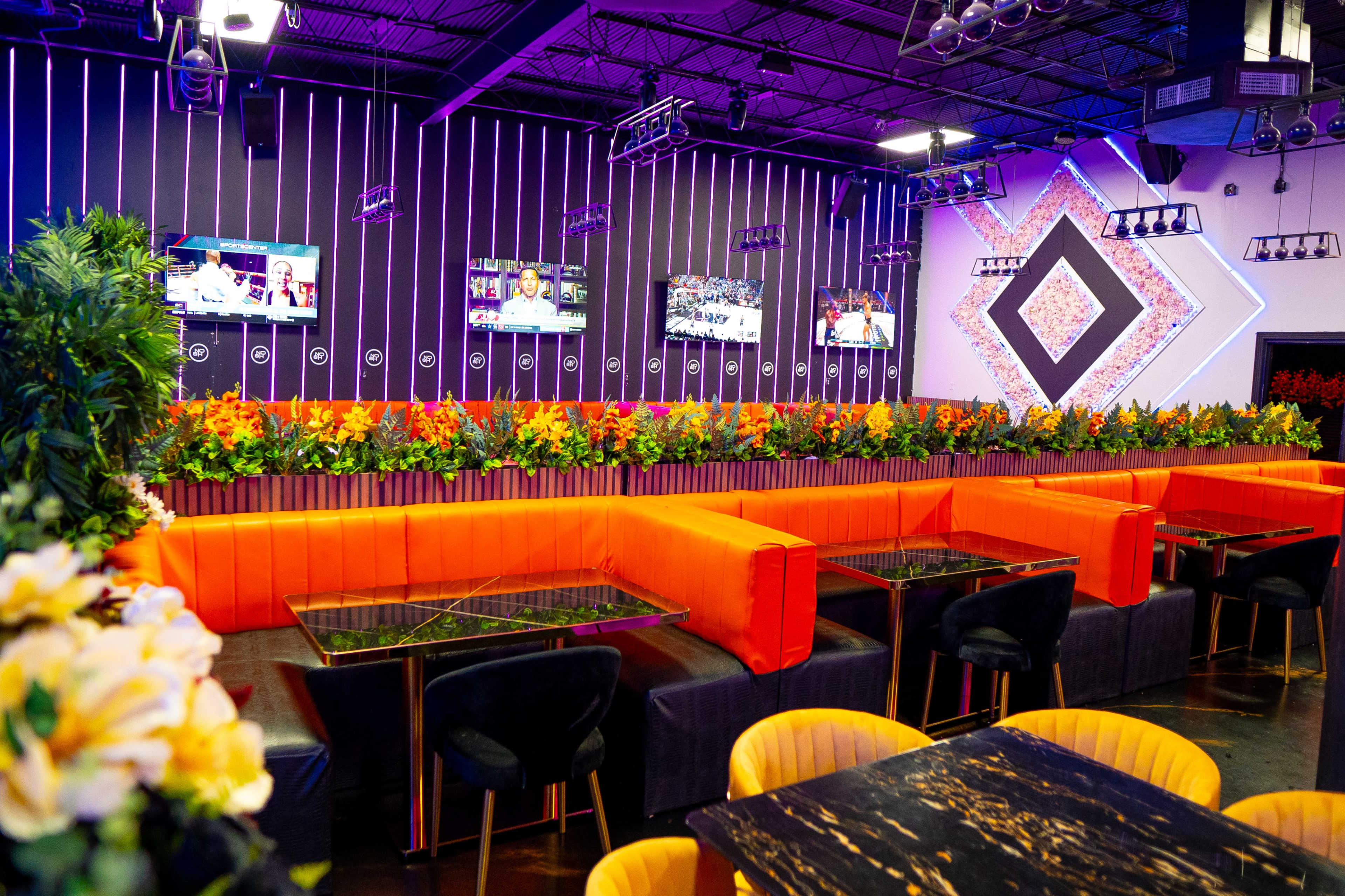 The image shows a modern lounge area featuring vibrant orange seating, floral decorations along the back wall, and multiple screens displaying content.