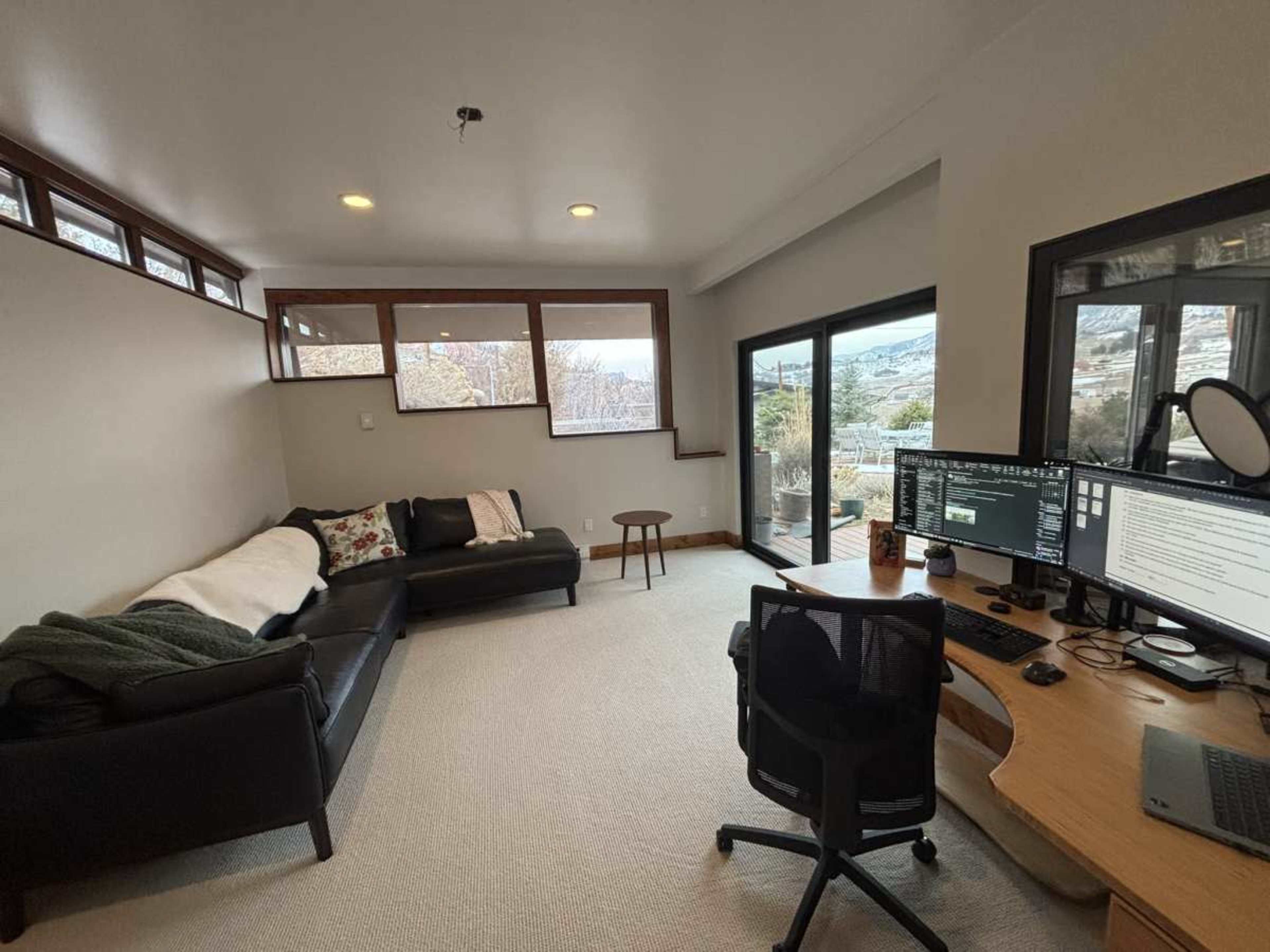 The image shows a tidy home office with a black leather couch, a wooden desk with dual monitors, and large windows that provide a view of the outdoors.