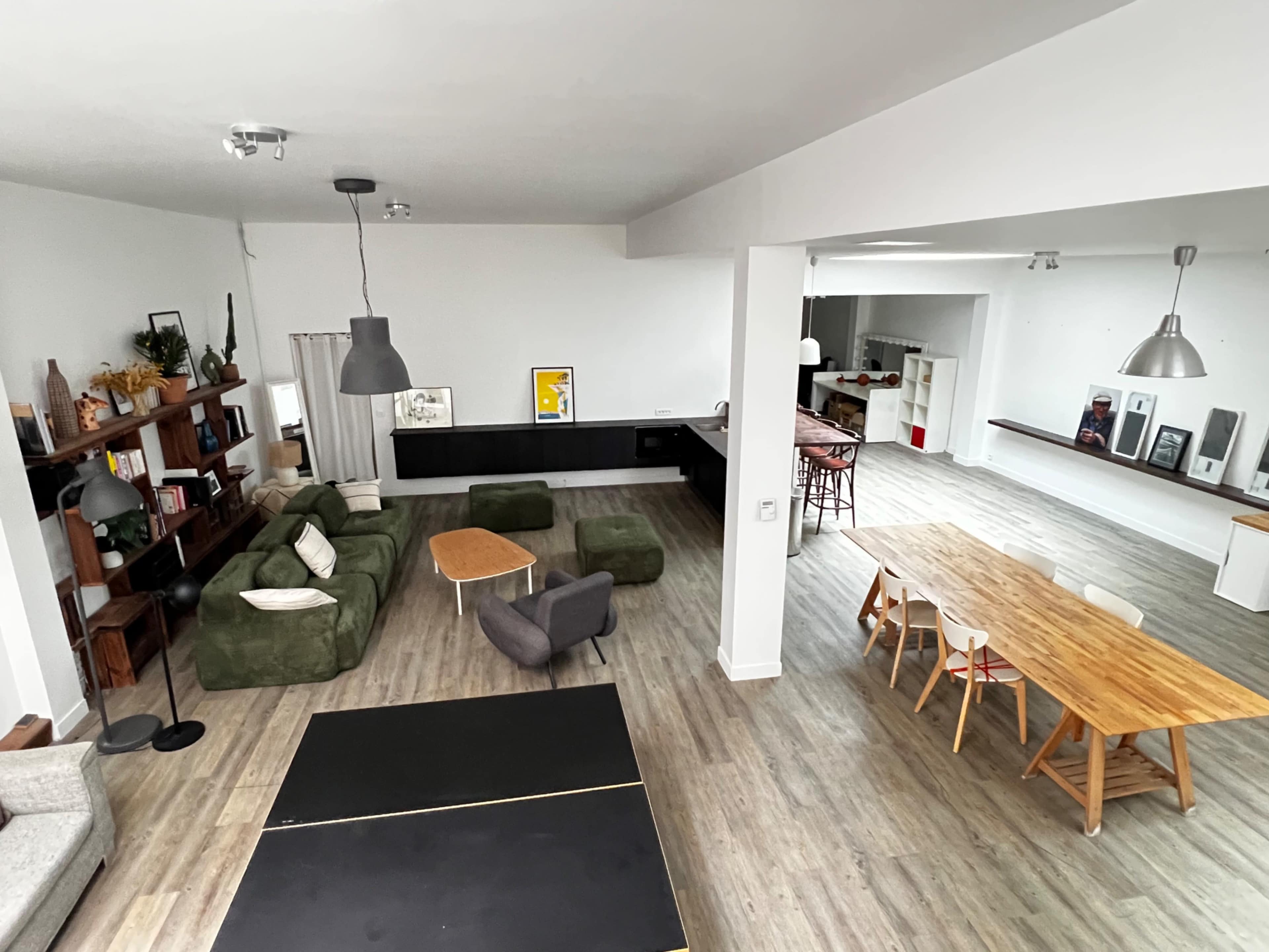 The image shows a spacious, modern living area featuring a green sectional sofa, a dining table with chairs, and a kitchen area in the background.