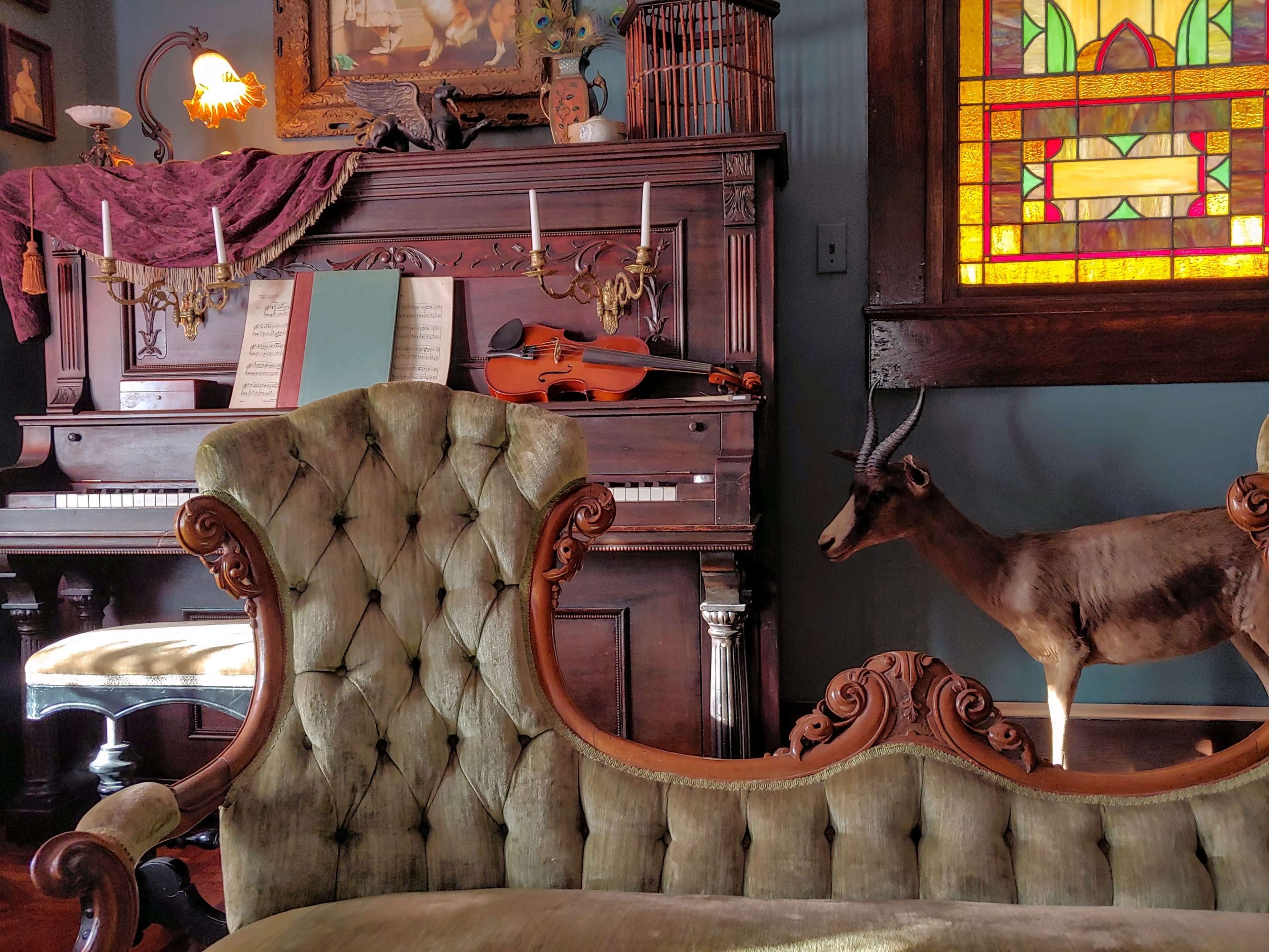 The image features a vintage room with a dark wooden piano, a violin, sheet music, a green tufted sofa, and a mounted deer on display near a stained glass window.