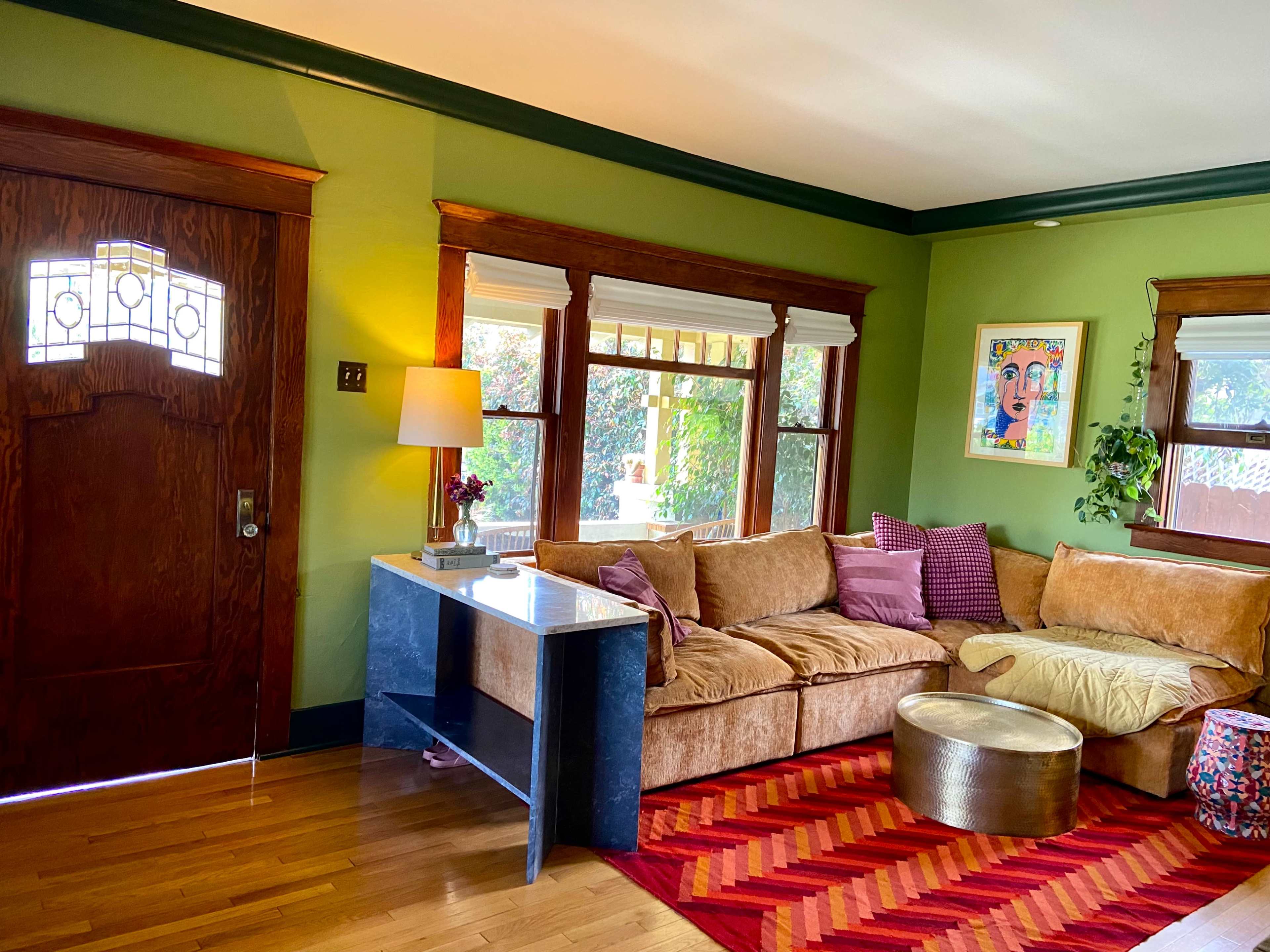 The image shows a cozy living room with a brown sectional sofa, a small marble-topped table, and vibrant green walls, illuminated by natural light from the windows.