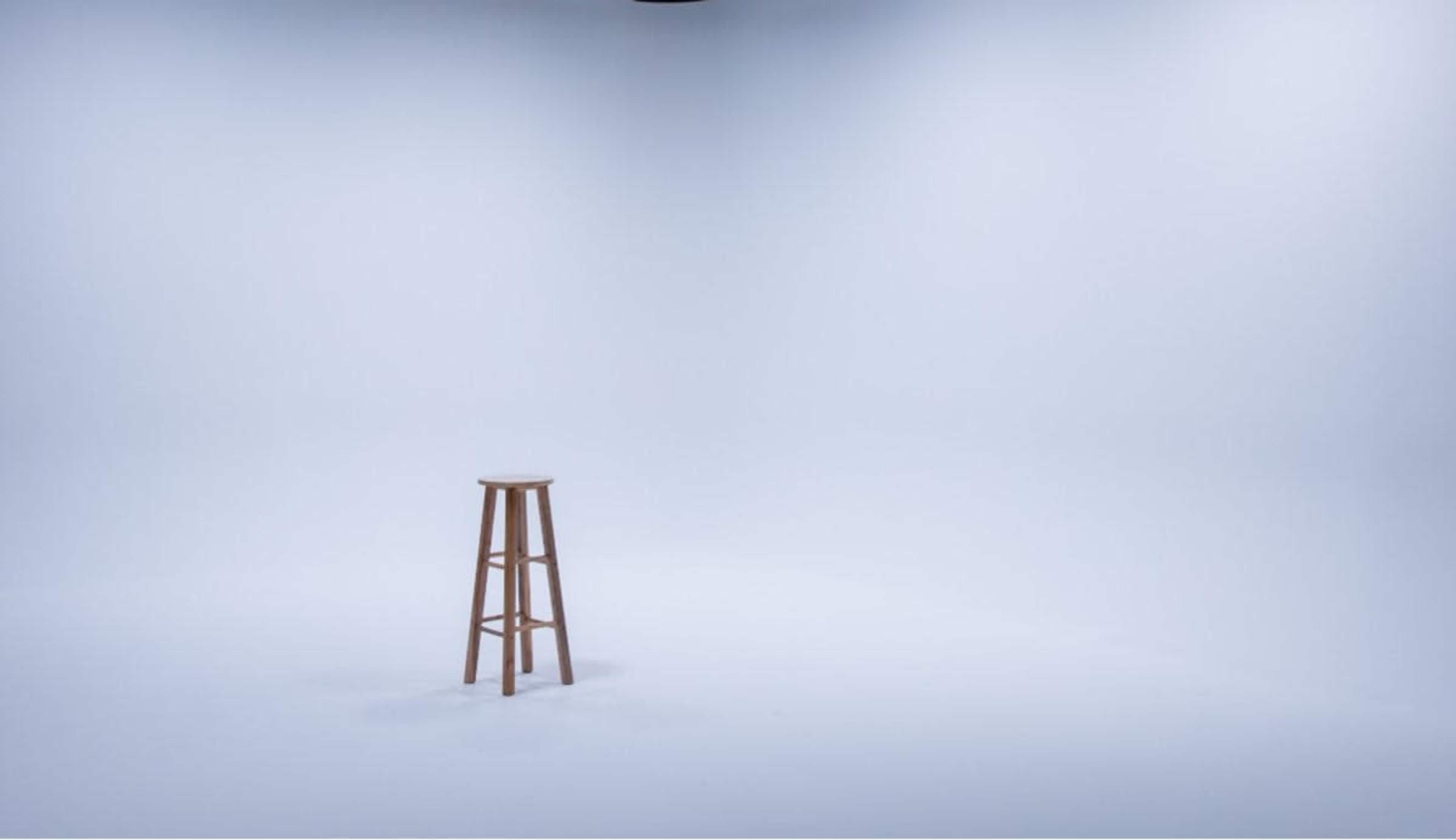 A wooden stool stands alone in a well-lit, empty studio with a light gray background.