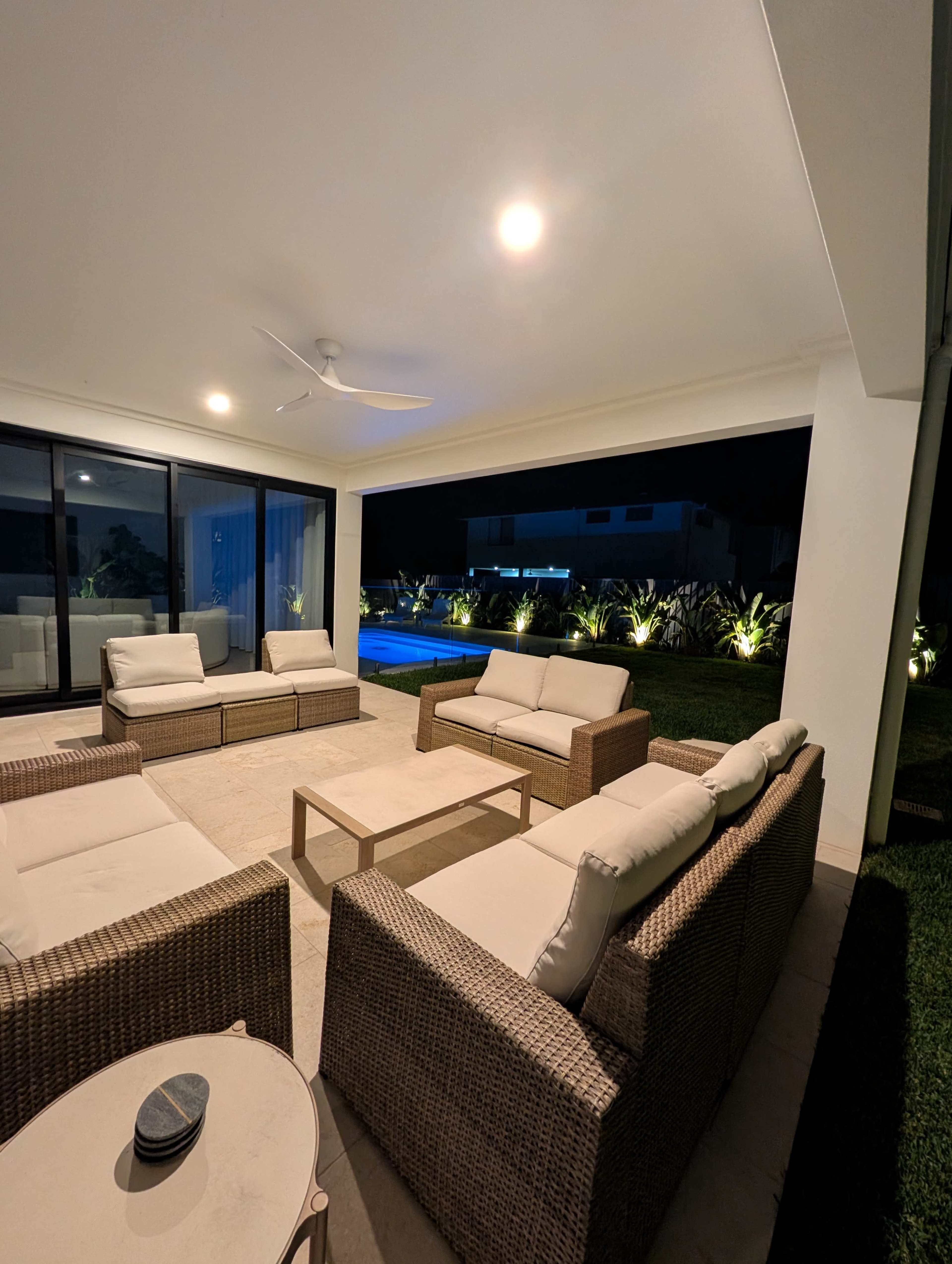 A modern outdoor seating area features multiple wicker sofas and a coffee table, illuminated by soft lighting, with a view of a well-lit pool and landscaped garden at night.