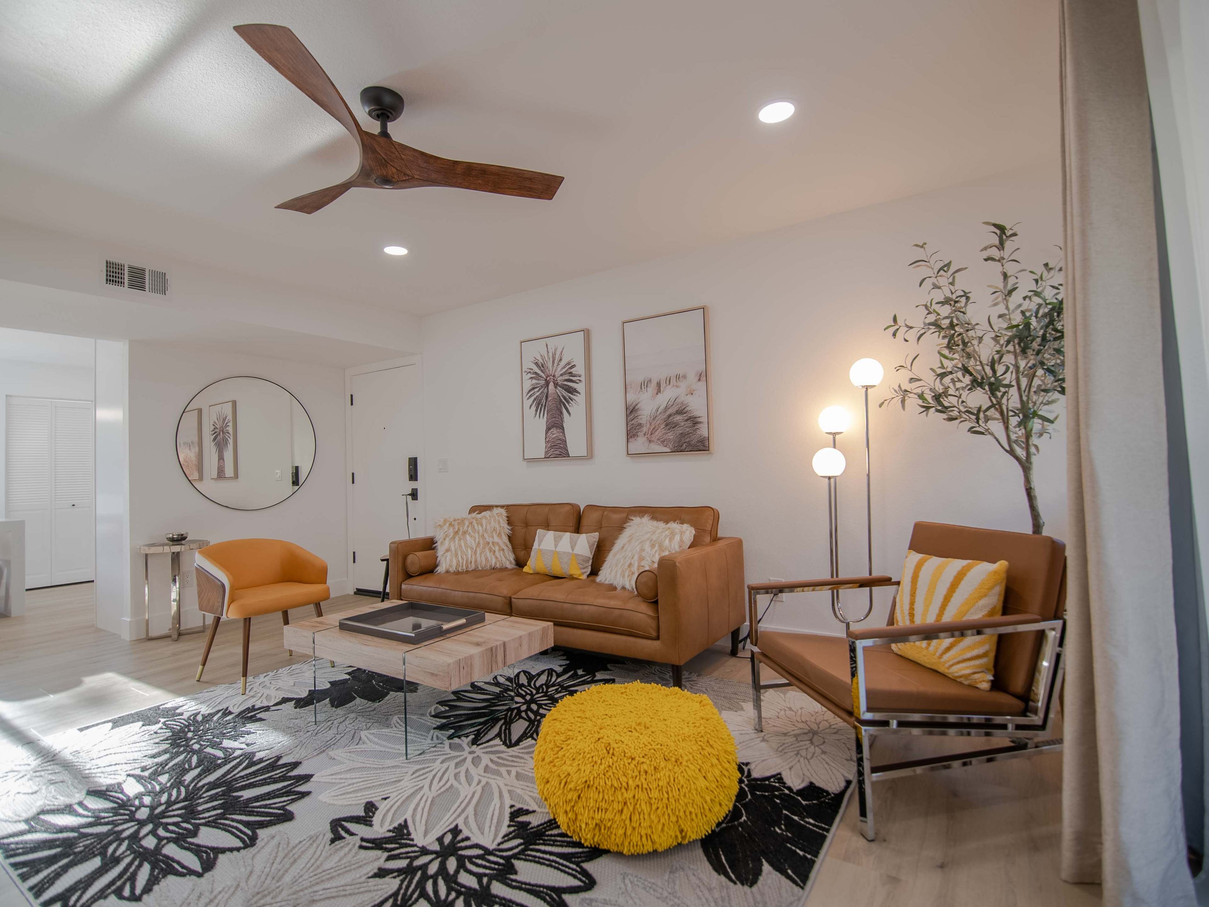 The living room features a brown sofa, an orange chair, a coffee table, and a large area rug with floral patterns, illuminated by overhead lighting and natural light.