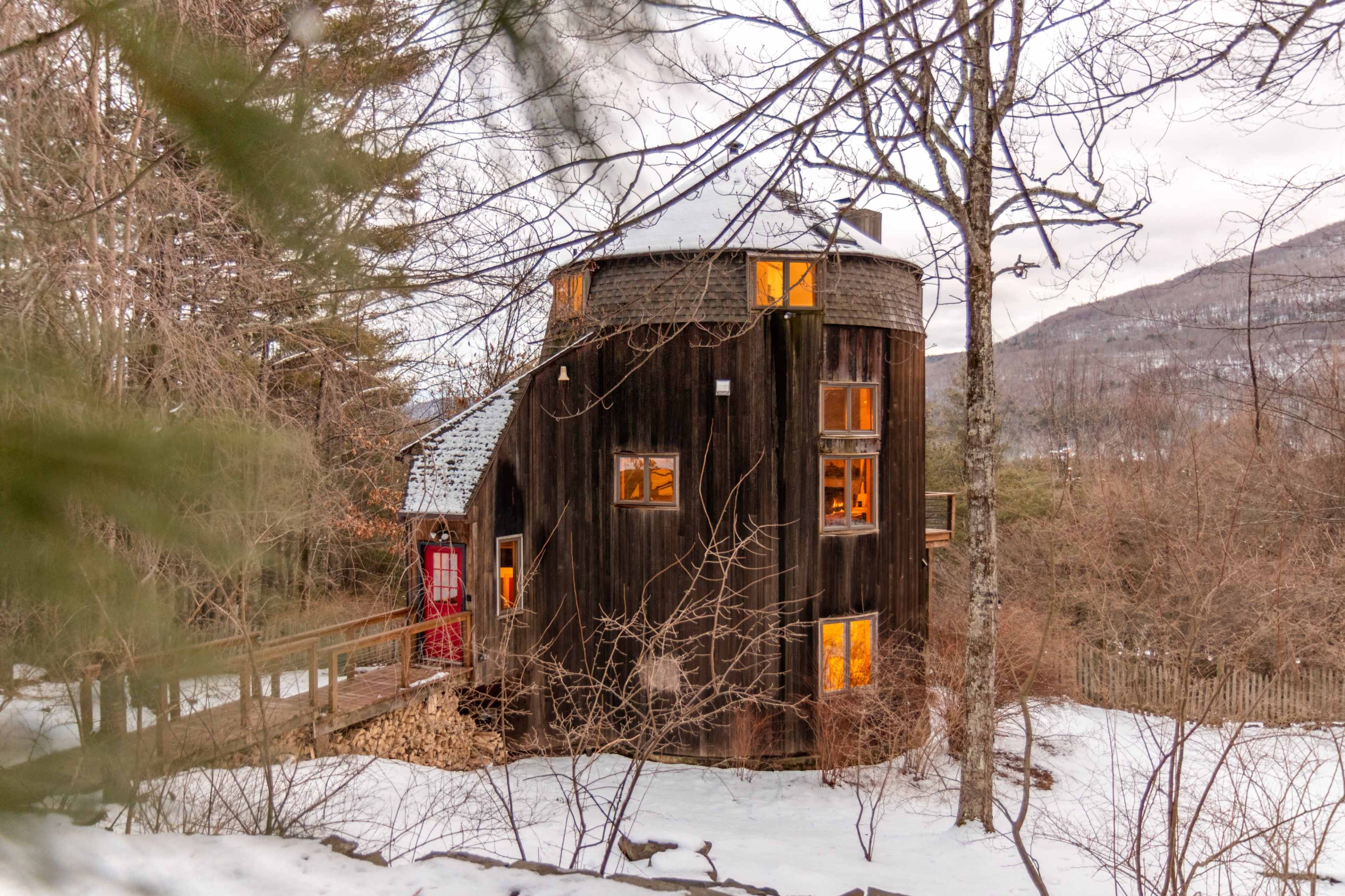 A round, wooden house with large windows is surrounded by trees and snow, set against a backdrop of hills.