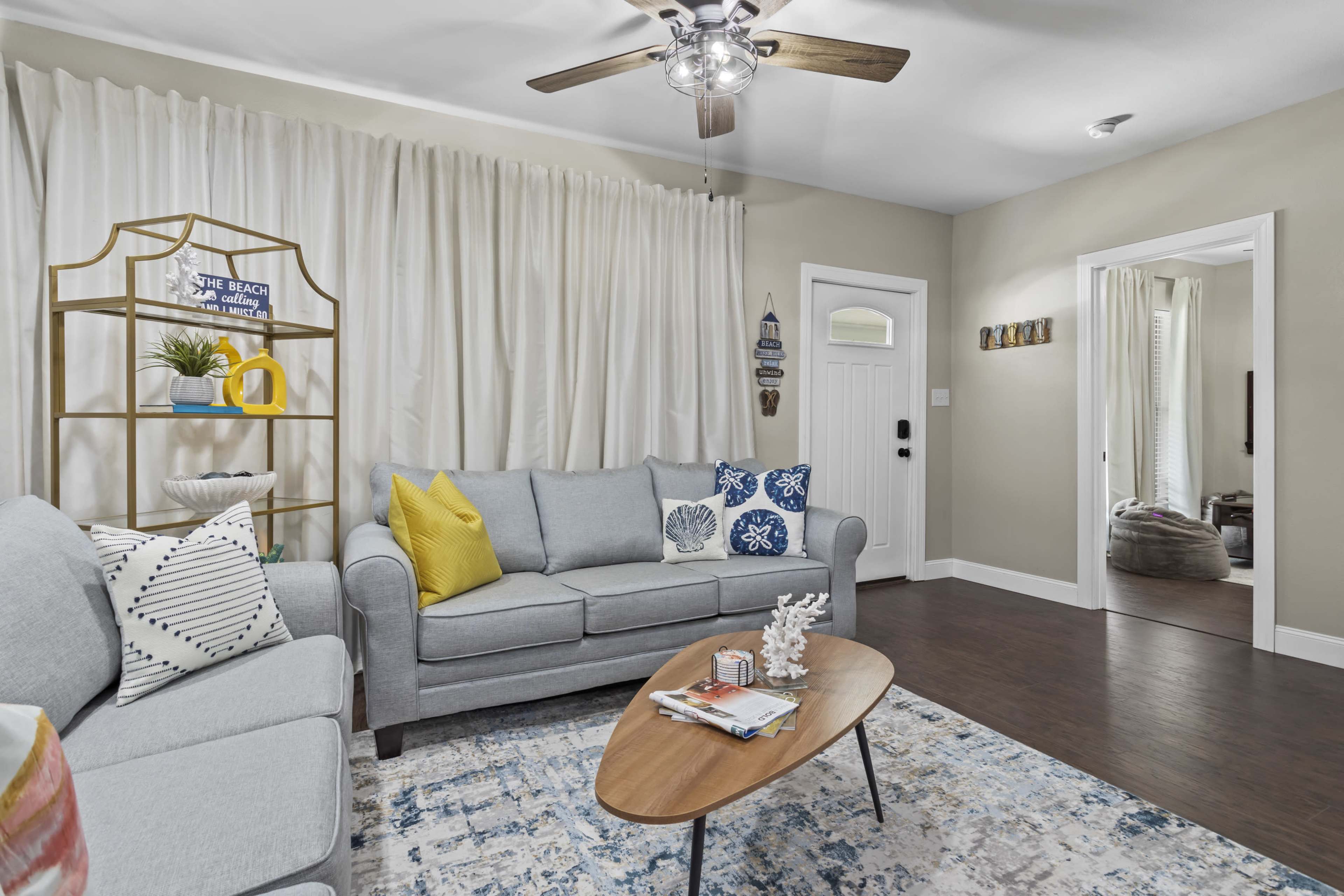 A living room features a light gray sofa with decorative pillows, a wooden coffee table, and a bookshelf against a wall with white curtains.