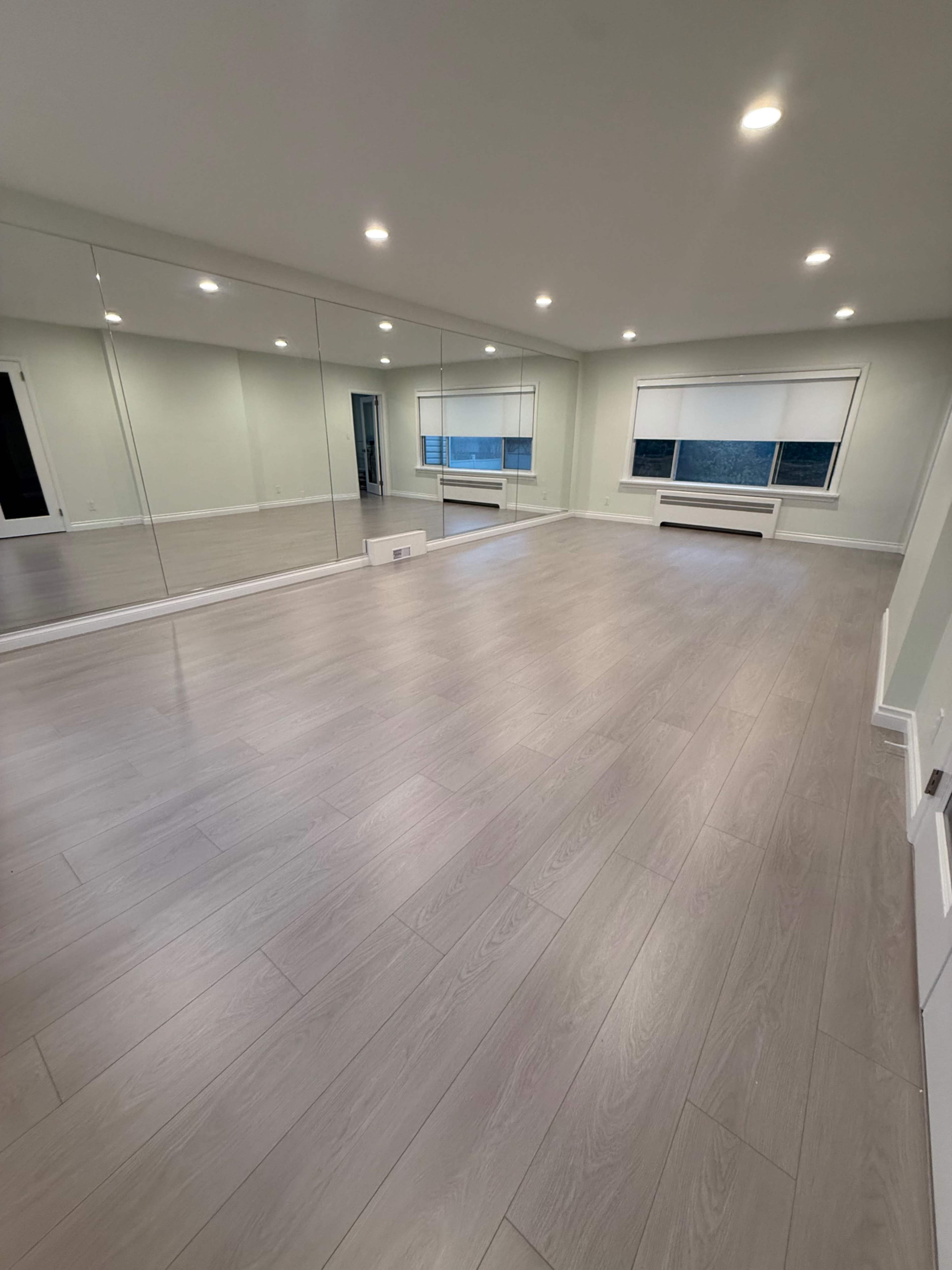 The room features light-colored laminate flooring, large mirrors on one wall, and a window with blinds, creating a spacious and airy environment.