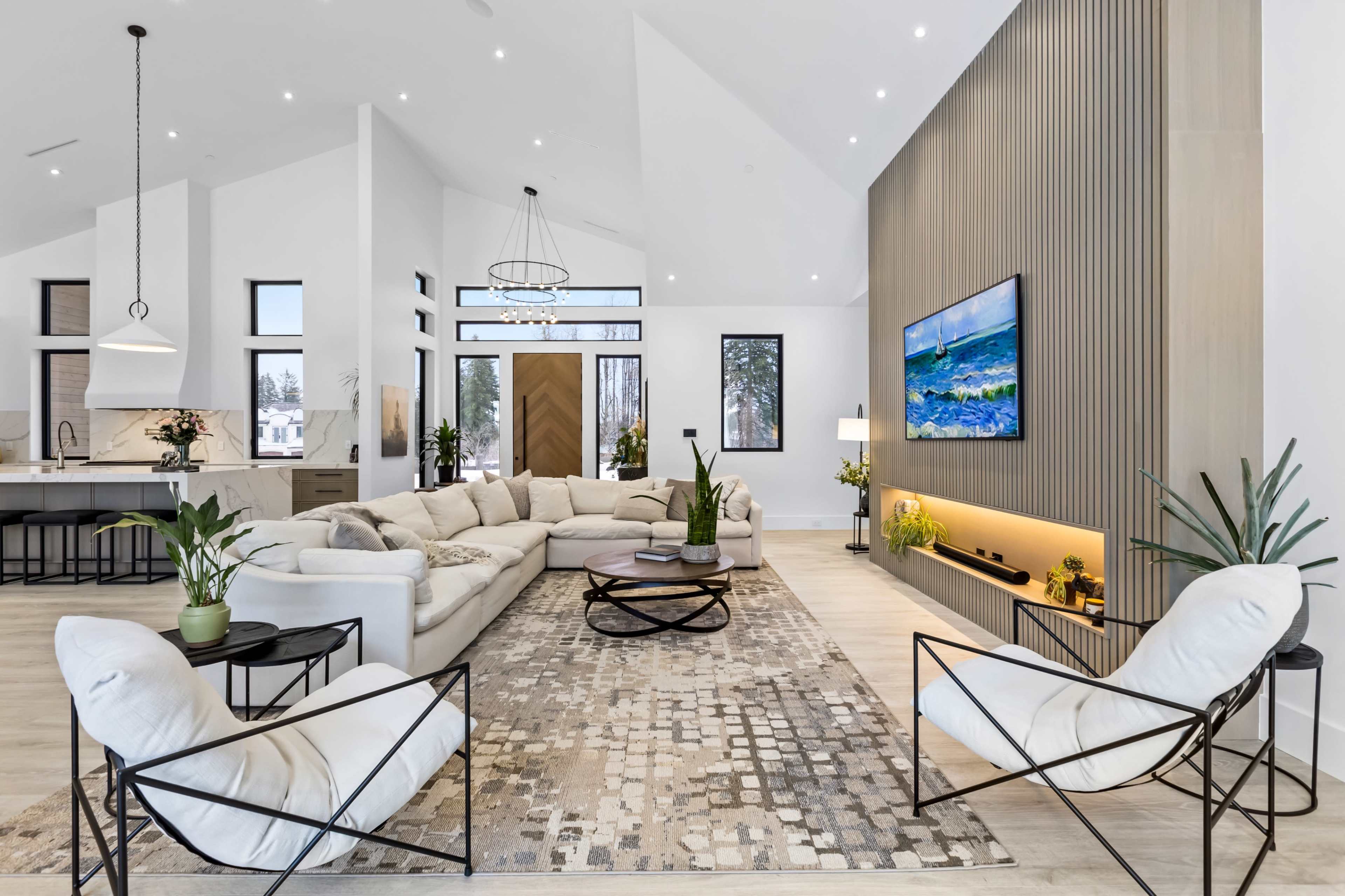 The image shows a modern living room with a sectional sofa, two black chairs, a coffee table, and a large wall-mounted TV, complemented by a decorative rug and plants.