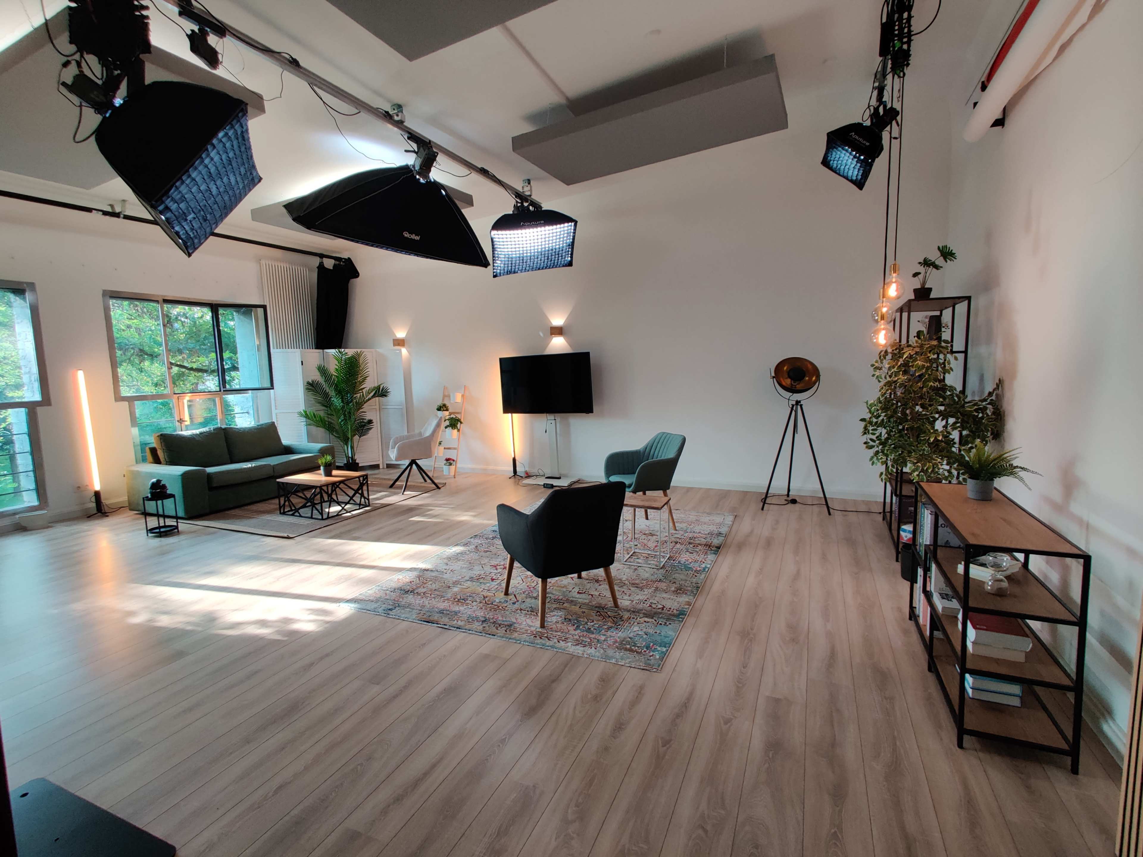 A well-lit studio space with a green sofa, two chairs, a large rug, and various decorative plants, along with studio lighting equipment and a television setup.