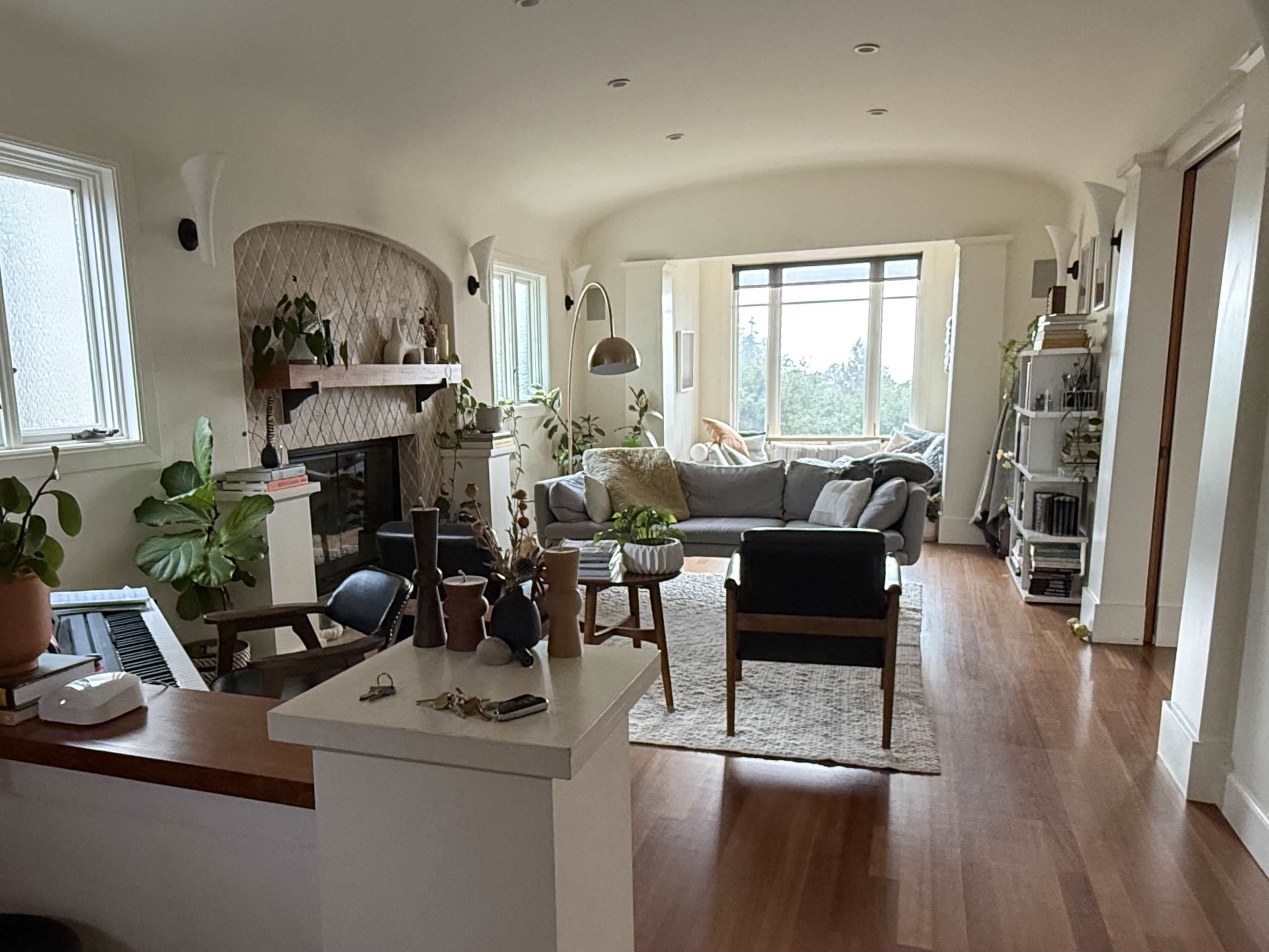 The image shows a spacious living room with a gray sofa, hardwood floors, and various plants, along with a fireplace and large windows allowing natural light.