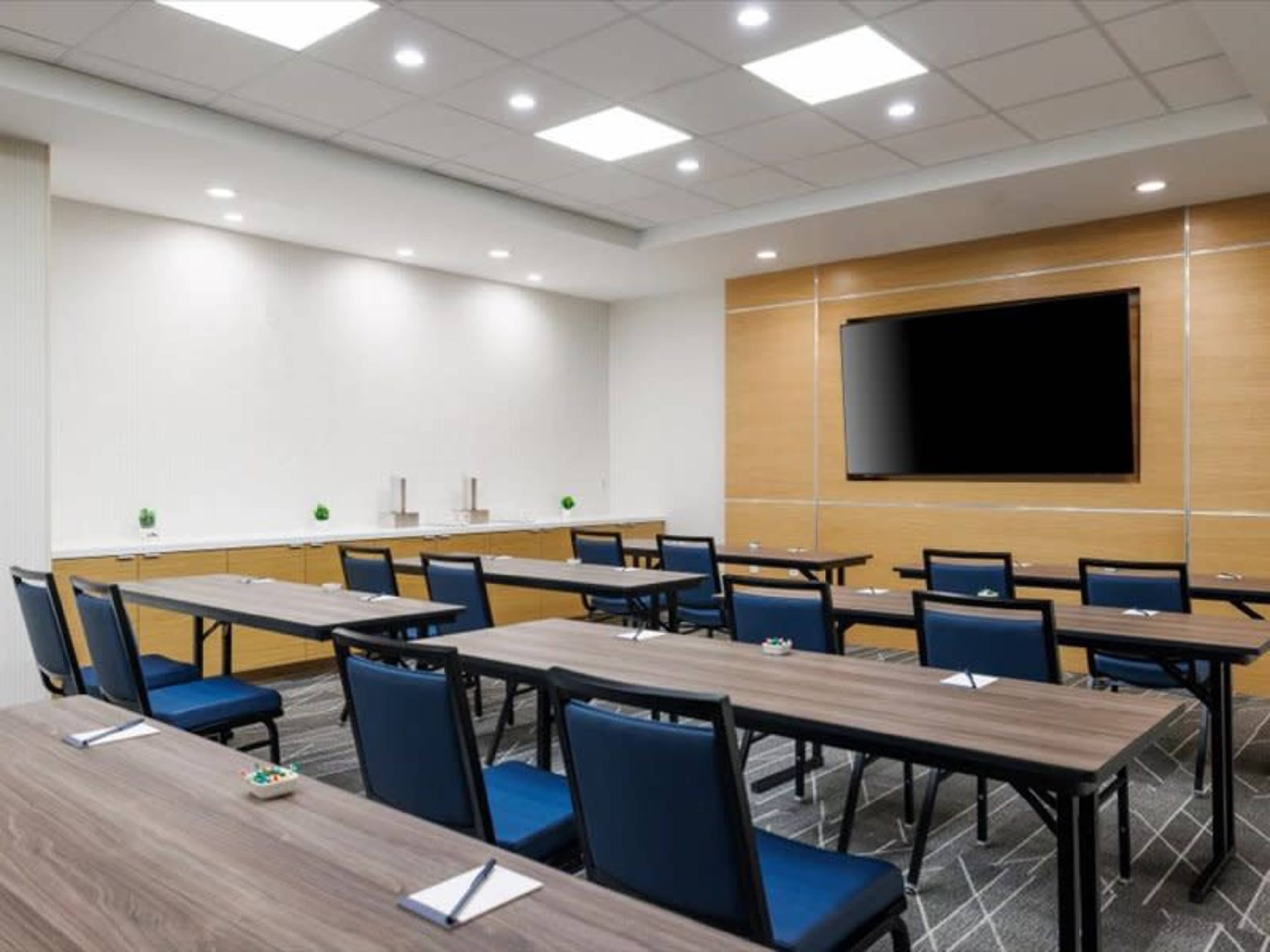 A conference room features several rows of tables and chairs arranged for a meeting, with a television mounted on the wall and light fixtures overhead.